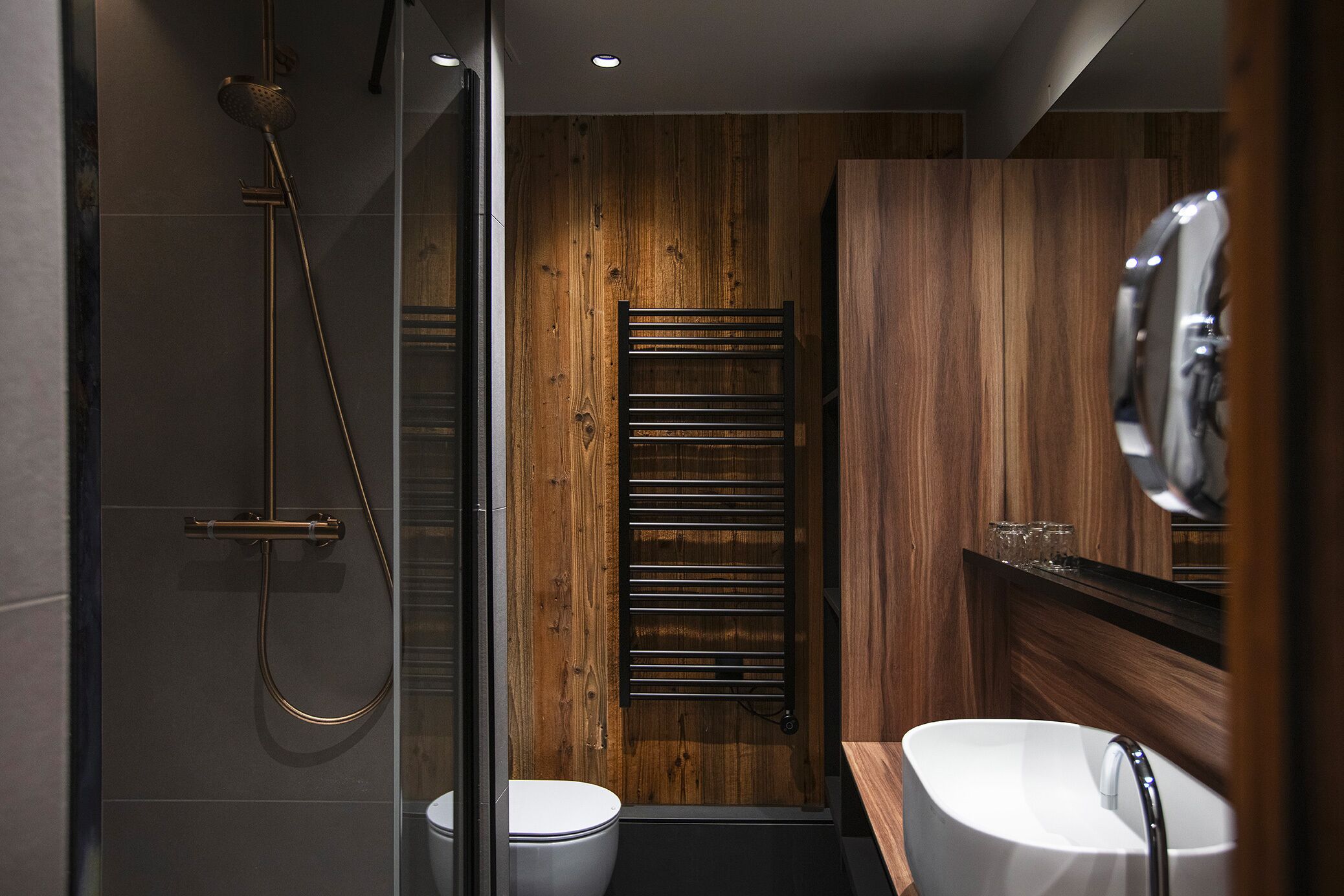 Un bagno moderno con doccia vetrata, pannello parete in legno, porta asciugamani e lavandino bianco con rubinetto cromato.