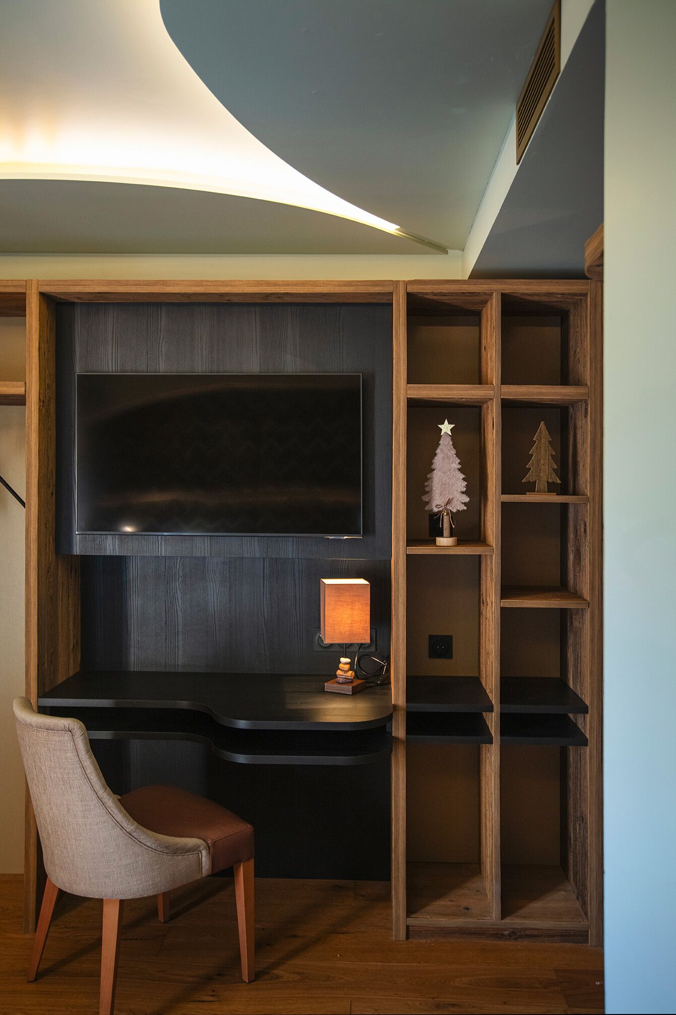 Un espacio de oficina moderno con una televisión de pantalla plana grande montada en la pared, un escritorio con una lámpara y una silla. Hay estanterías de madera con objetos decorativos.