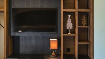 Un espacio de oficina moderno con una televisión de pantalla plana grande montada en la pared, un escritorio con una lámpara y una silla. Hay estanterías de madera con objetos decorativos.