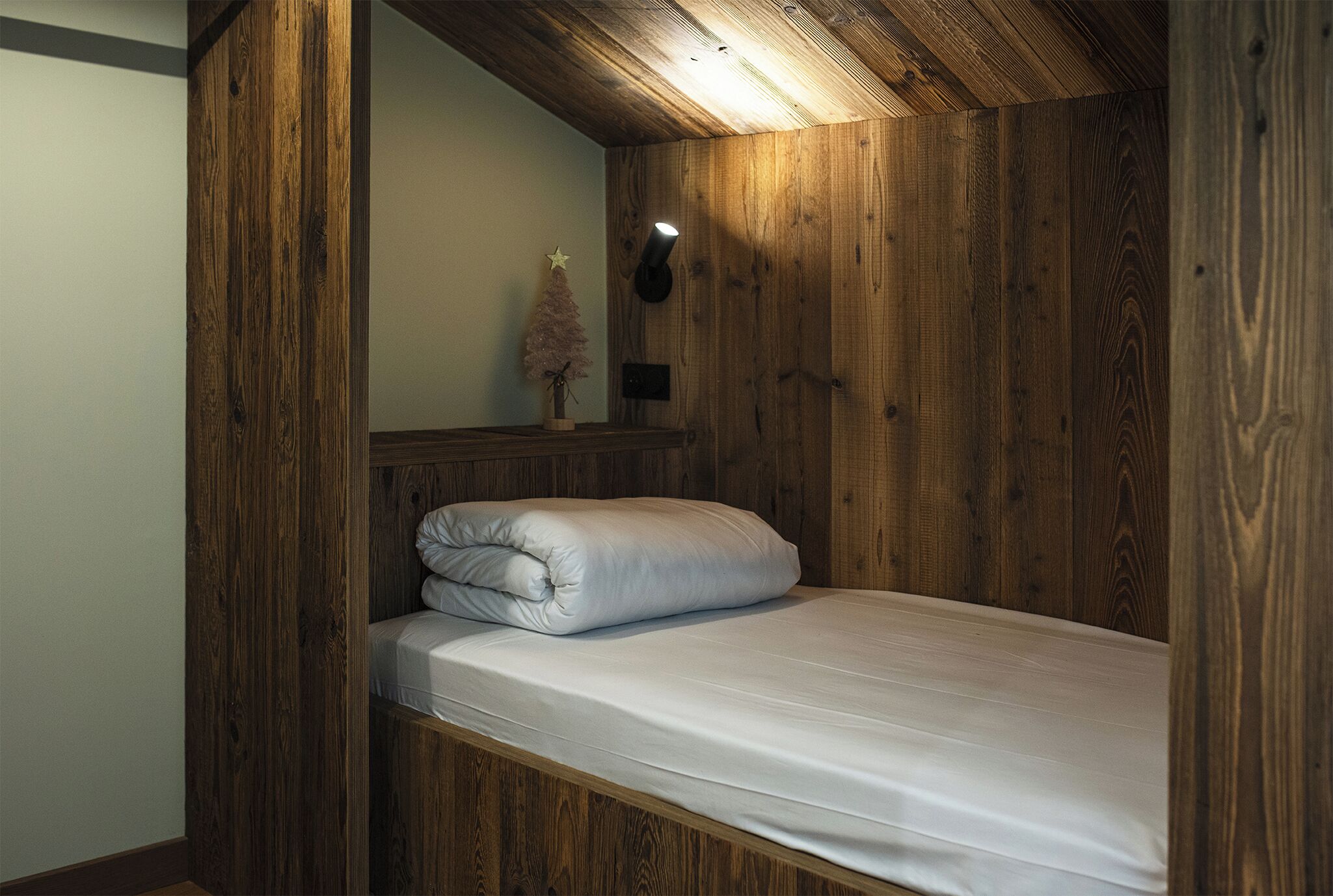 Una cama litera de madera con colchón y almohadas blancas, un pequeño árbol de Navidad y un aplique de pared.