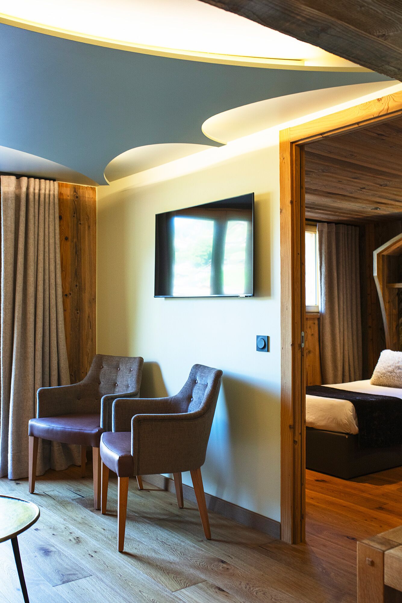 Una habitación con una cama, dos sillones, un televisor montado en la pared y suelo de madera.