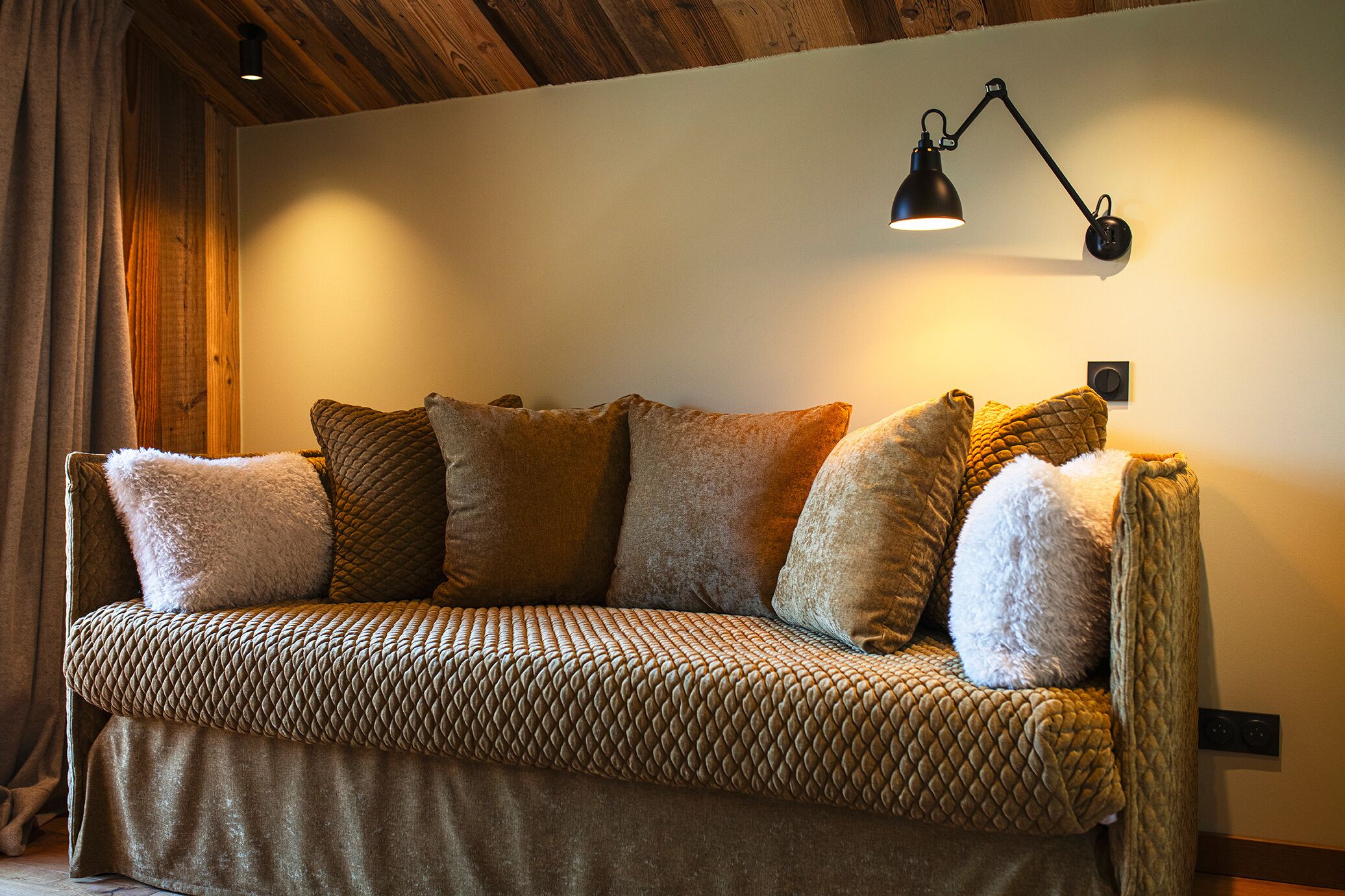 A cozy sofa with a quilted pattern and multiple cushions, a wall-mounted lamp, and wooden ceiling beams.