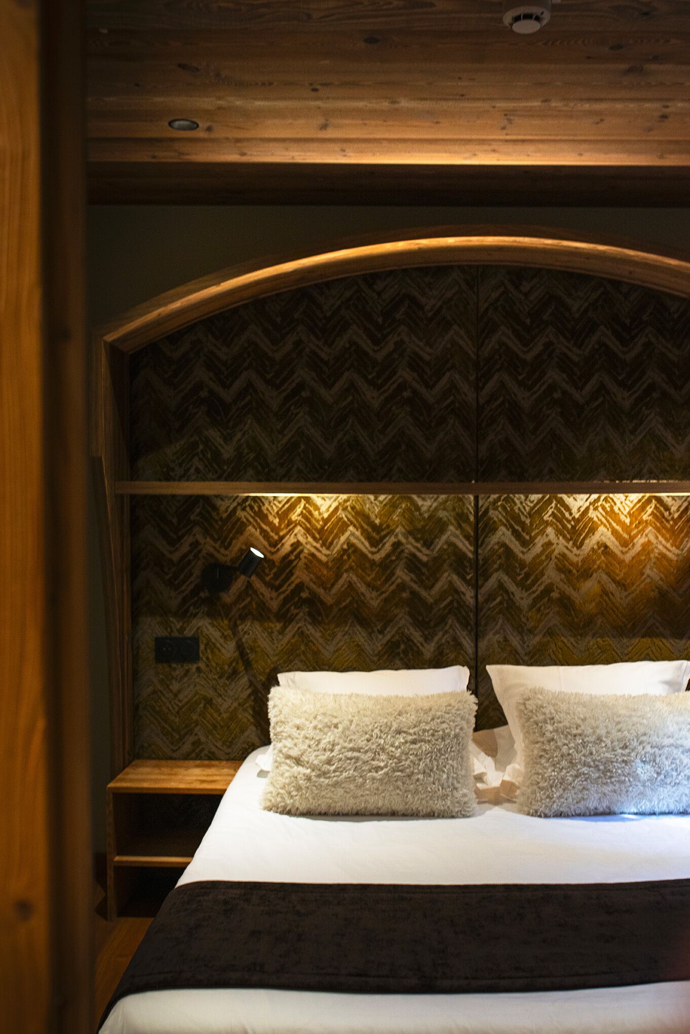 A hotel room with a bed, wooden headboard, and a herringbone-patterned wall.