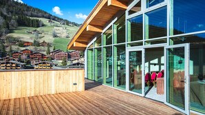 Un edificio moderno con grandes ventanales, una terraza de madera y vistas a un pueblo montañoso.