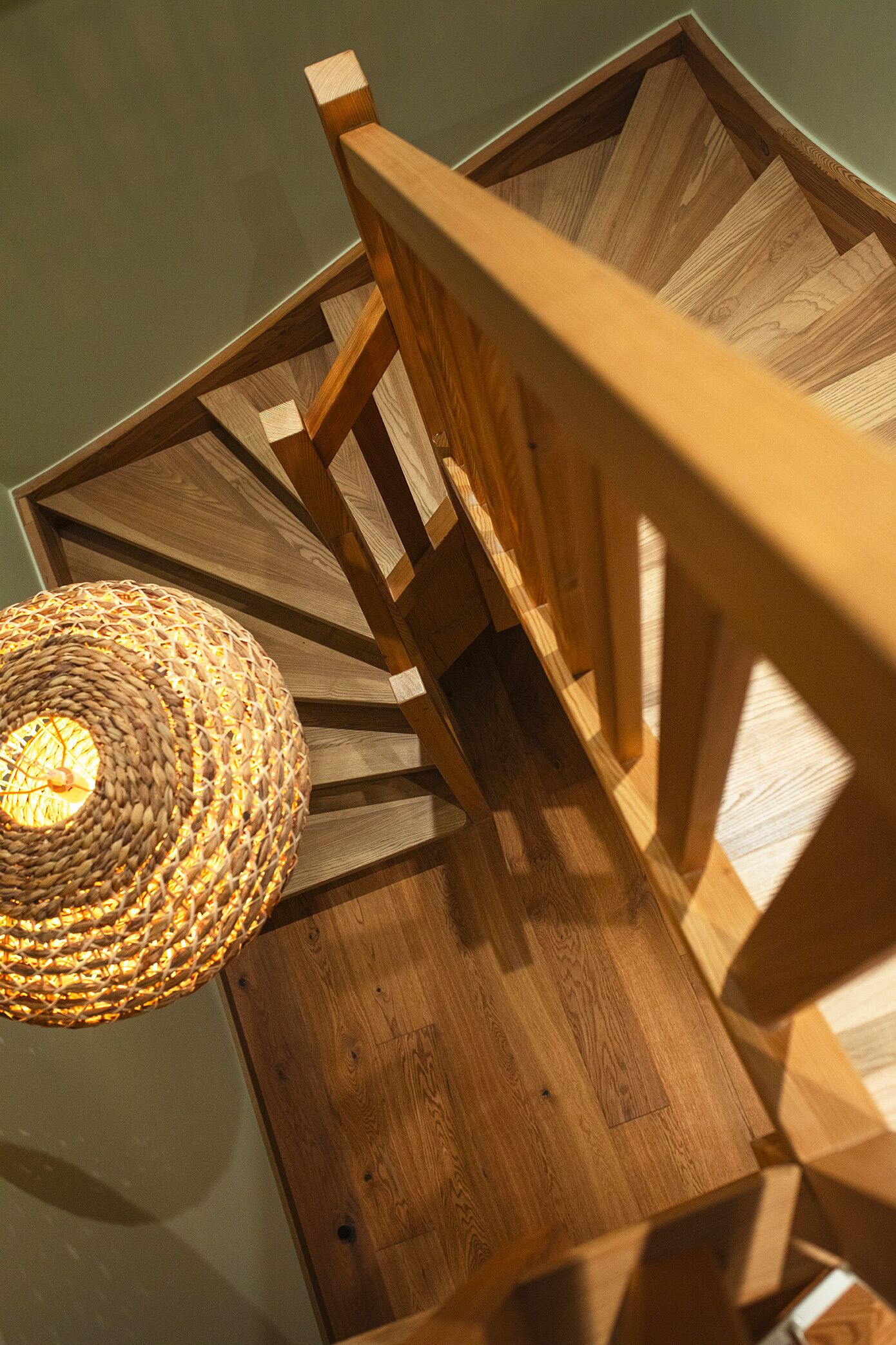 Una escalera en espiral de madera con una lámpara de fibra trenzada en la parte superior.