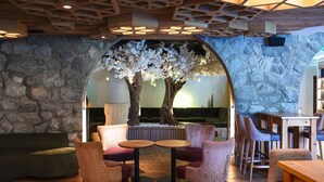 Un rincón interior acogedor con pared de piedra, techo de madera y un gran árbol con flores blancas. Hay mesas redondas y sillas con cojines.