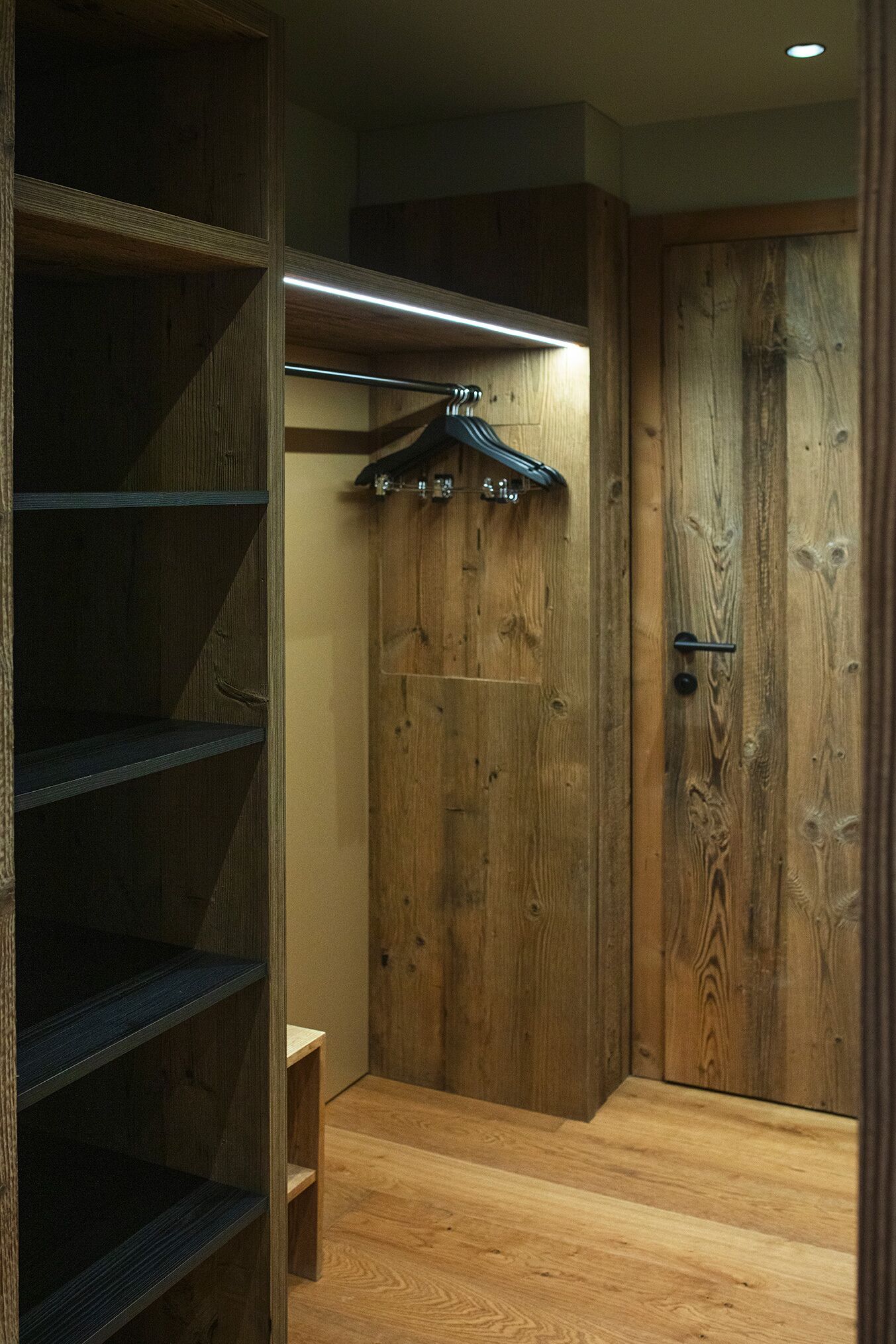 A wooden closet with shelves and a hanging rod, a wooden door, and a small bench.