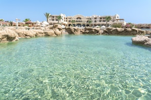 Une piscine aux eaux limpides, bordée de rochers, entourée d’un bâtiment d’hôtel doté de nombreux balcons et d’un palmier.