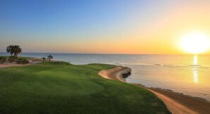 Um campo de golfe com uma zona de areia, palmeiras e vista para o oceano ao pôr do sol.