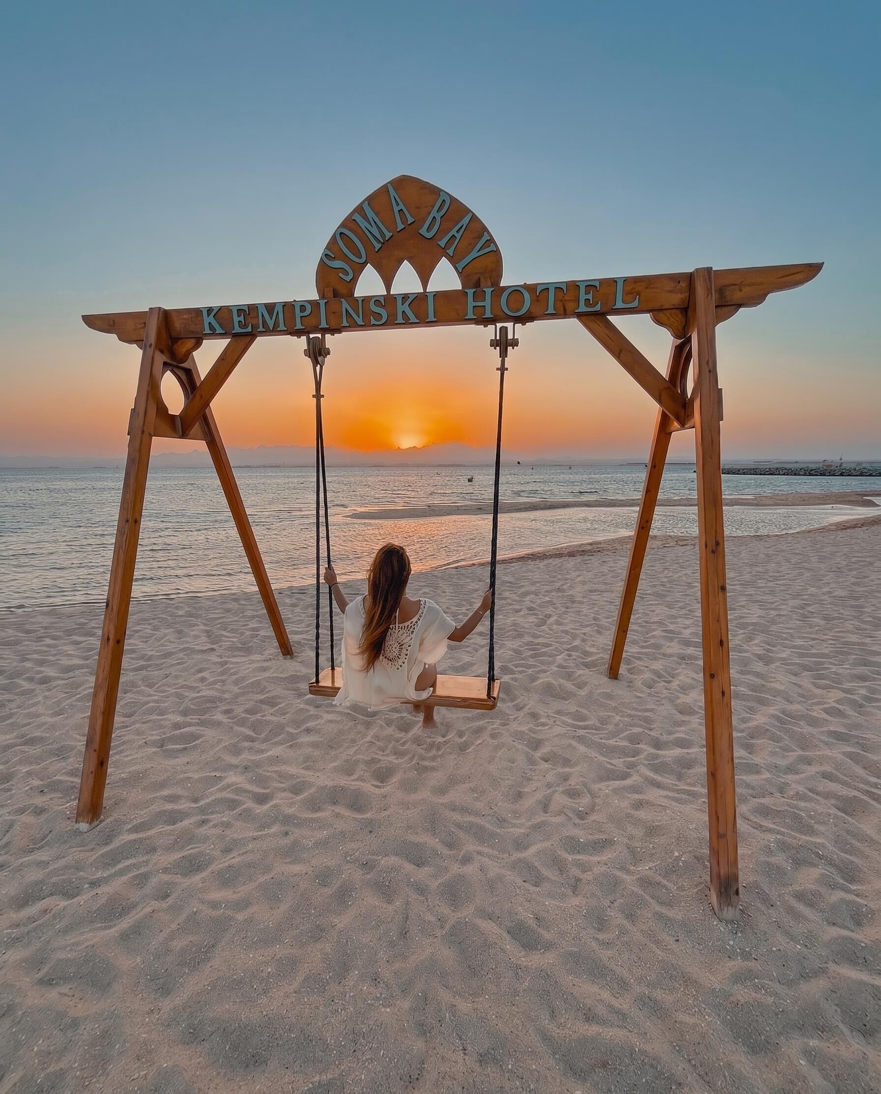 Eine Holz-Schaukel mit einem Schild, auf dem 'SOMABAY KEMPINSKI HOTEL' steht, an einem Strand bei Sonnenuntergang.