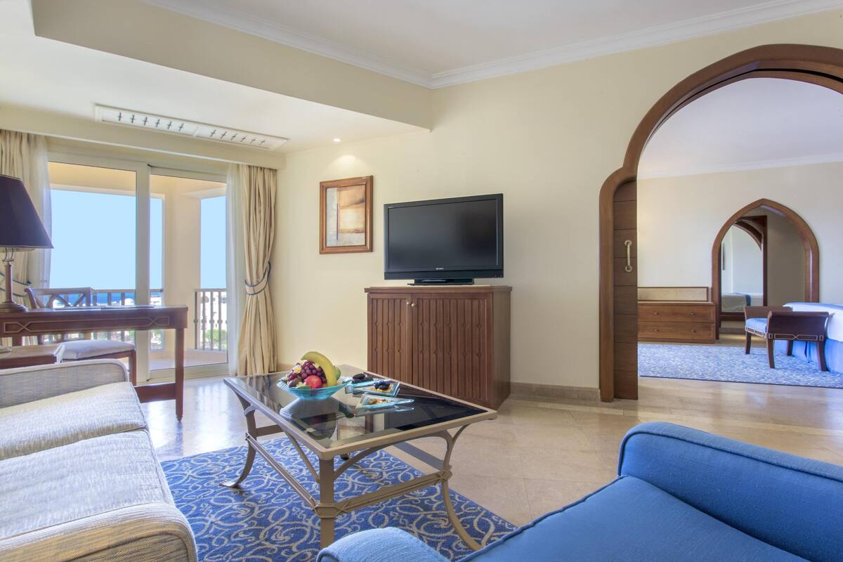 Ein Hotelzimmer mit Wohnbereich, einem Fernseher, einem Esstisch mit Obstschale und einem Balkon mit Vorhängen.