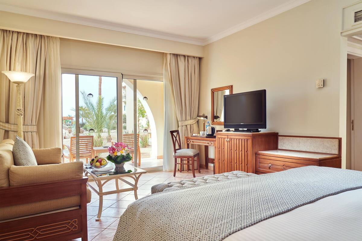 Ein Hotelzimmer mit einem großen Bett, einem Schreibtisch mit Fernseher, einem Sessel und einem Esstisch mit Obstschale.