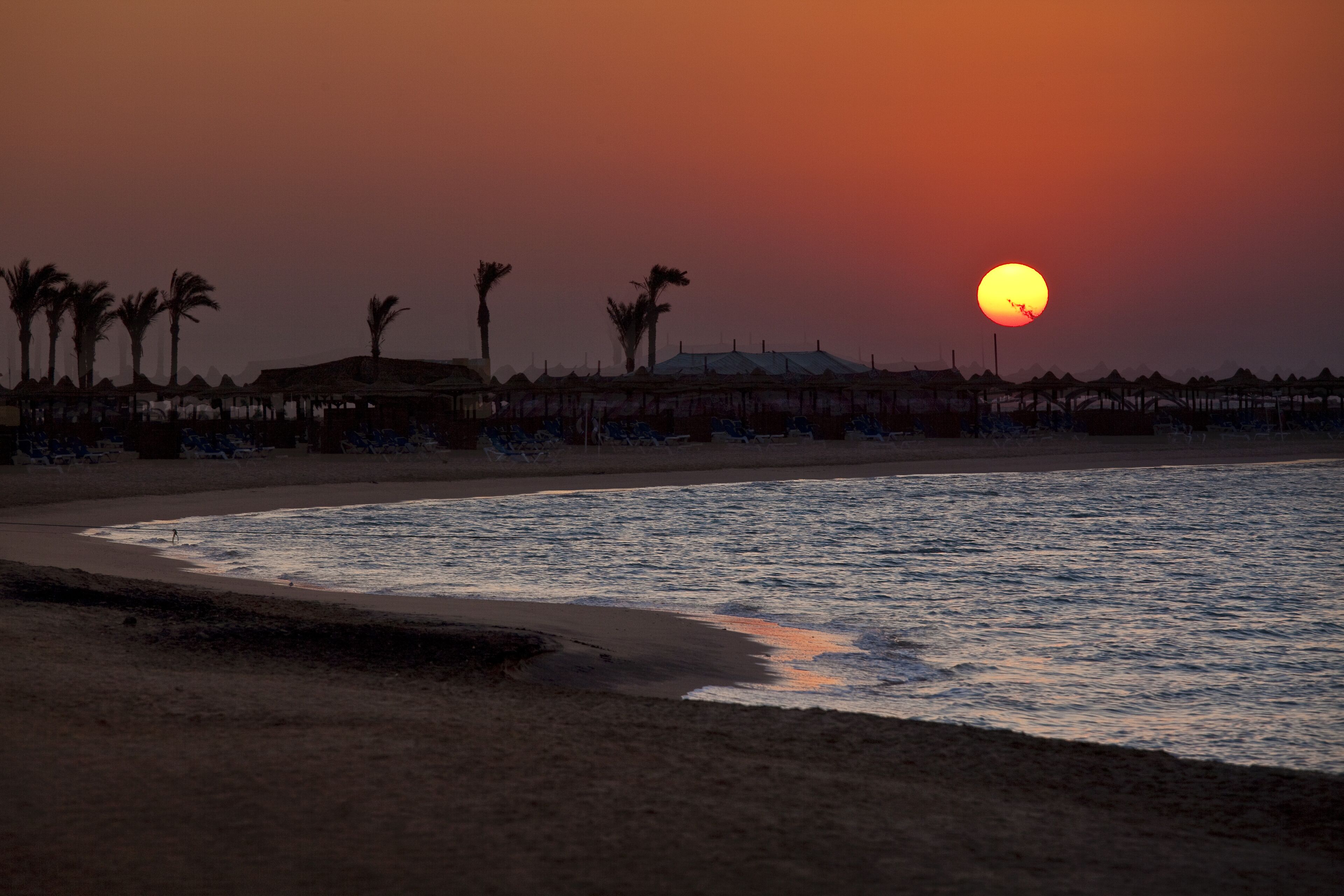 A beach with a sunset, palm trees, and beach huts.