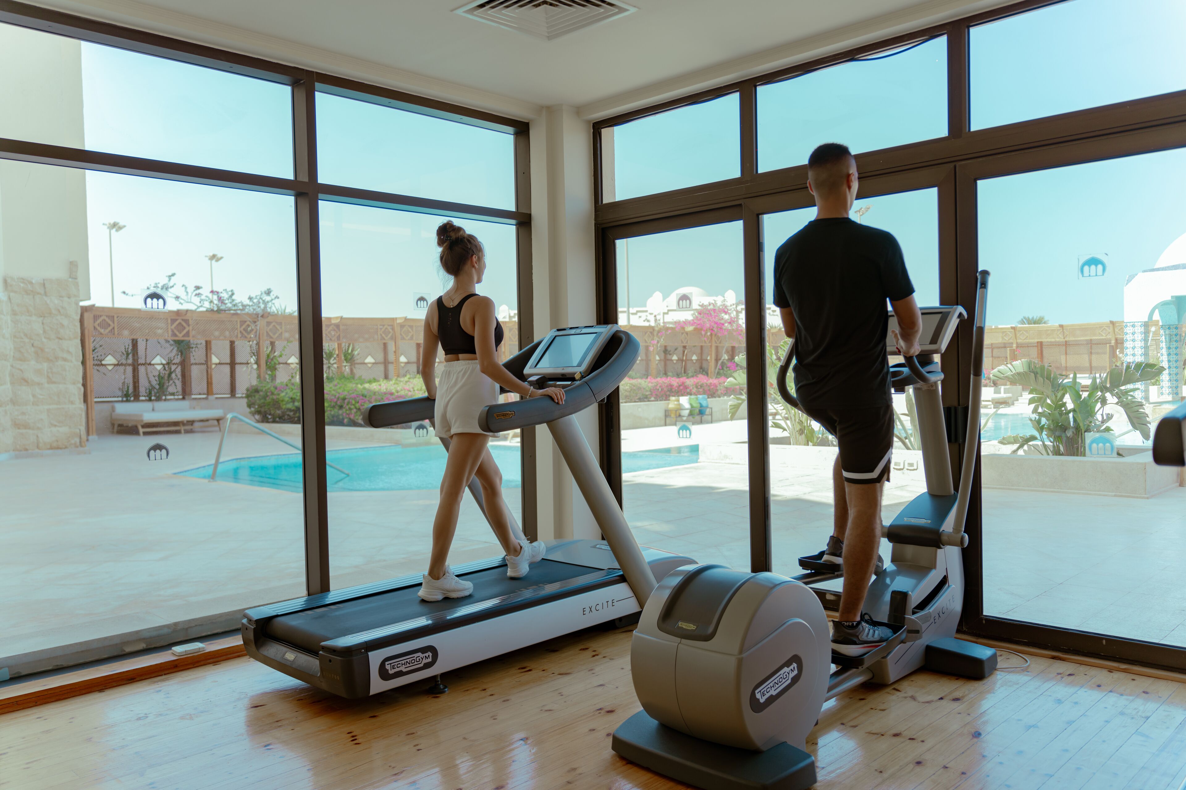 A gym with treadmills and a pool visible through large windows.