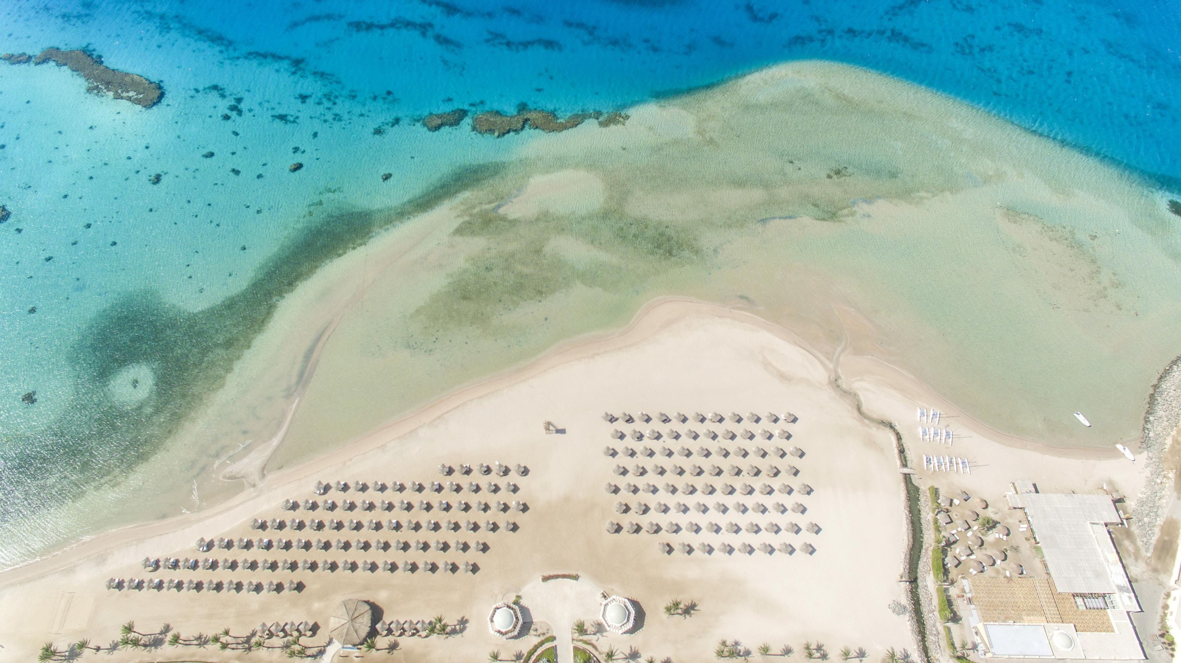 Vista aérea de un complejo turístico en la playa, con filas de tumbonas y sombrillas, una piscina y un pequeño edificio.