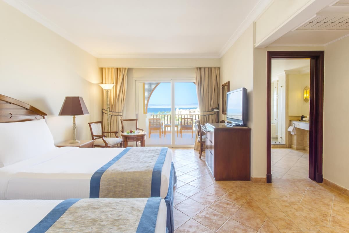 Ein Hotelzimmer mit zwei Betten, einem Fernseher, einem Schreibtisch und einem Balkon mit Meerblick.