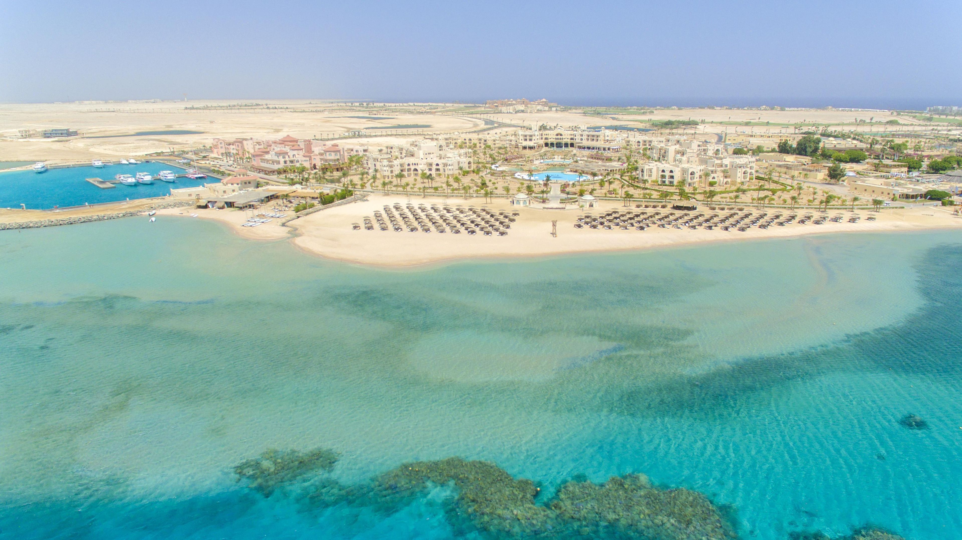 Vista aérea de un complejo turístico costero con una playa de arena, un puerto deportivo y un gran hotel.