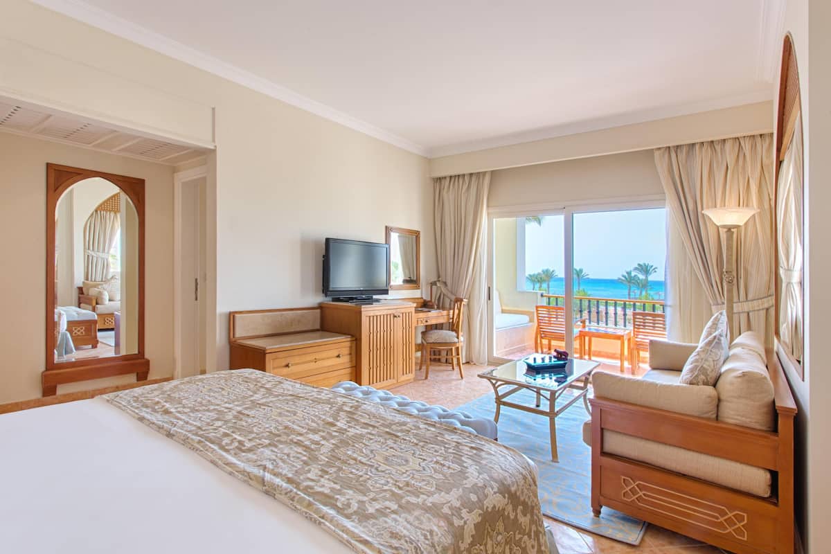 Ein Hotelzimmer mit einem großen Bett, einem Schreibtisch mit Fernseher, einem Balkon mit Meerblick und einem Sofa.