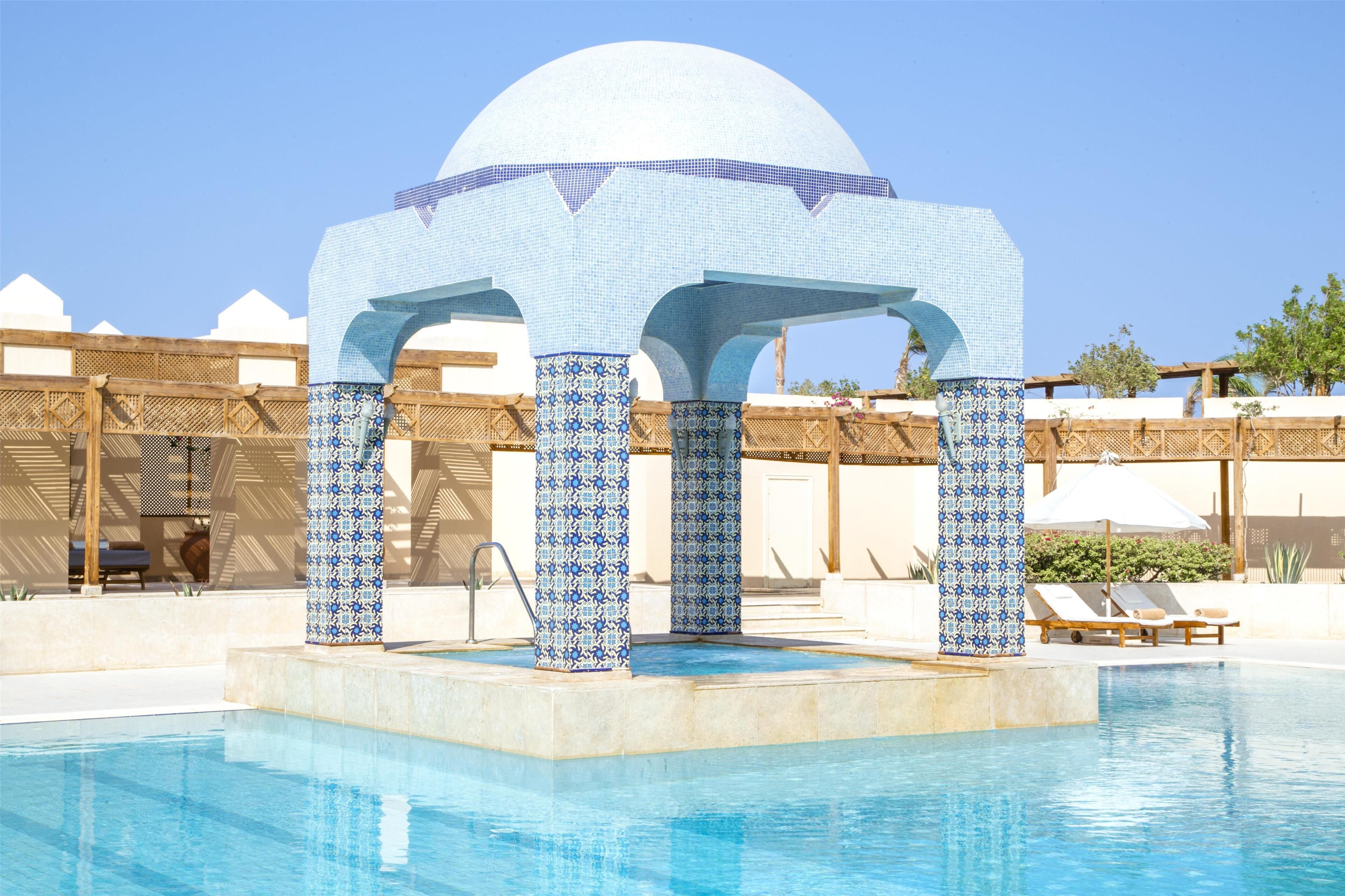A pool area with a tiled gazebo structure, lounge chairs, and a clear blue sky.