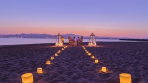 A beach setting with a table and chairs arranged for a meal, surrounded by candles.