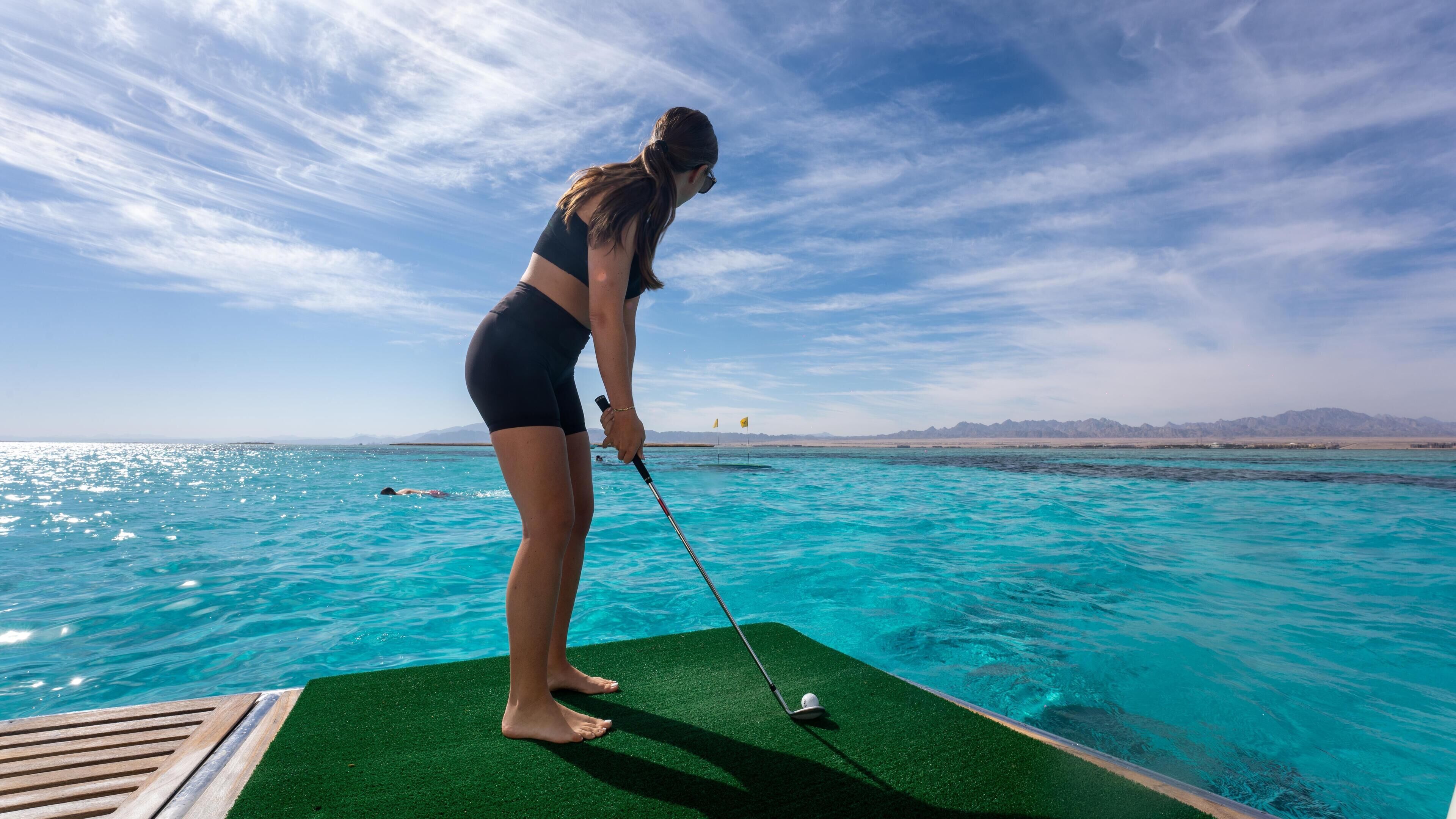 Una persona juega al golf sobre una plataforma verde junto a una masa de agua, con montañas al fondo.