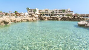 A resort with a clear pool surrounded by rocks and palm trees, a large building with multiple balconies, and a clear blue sky.