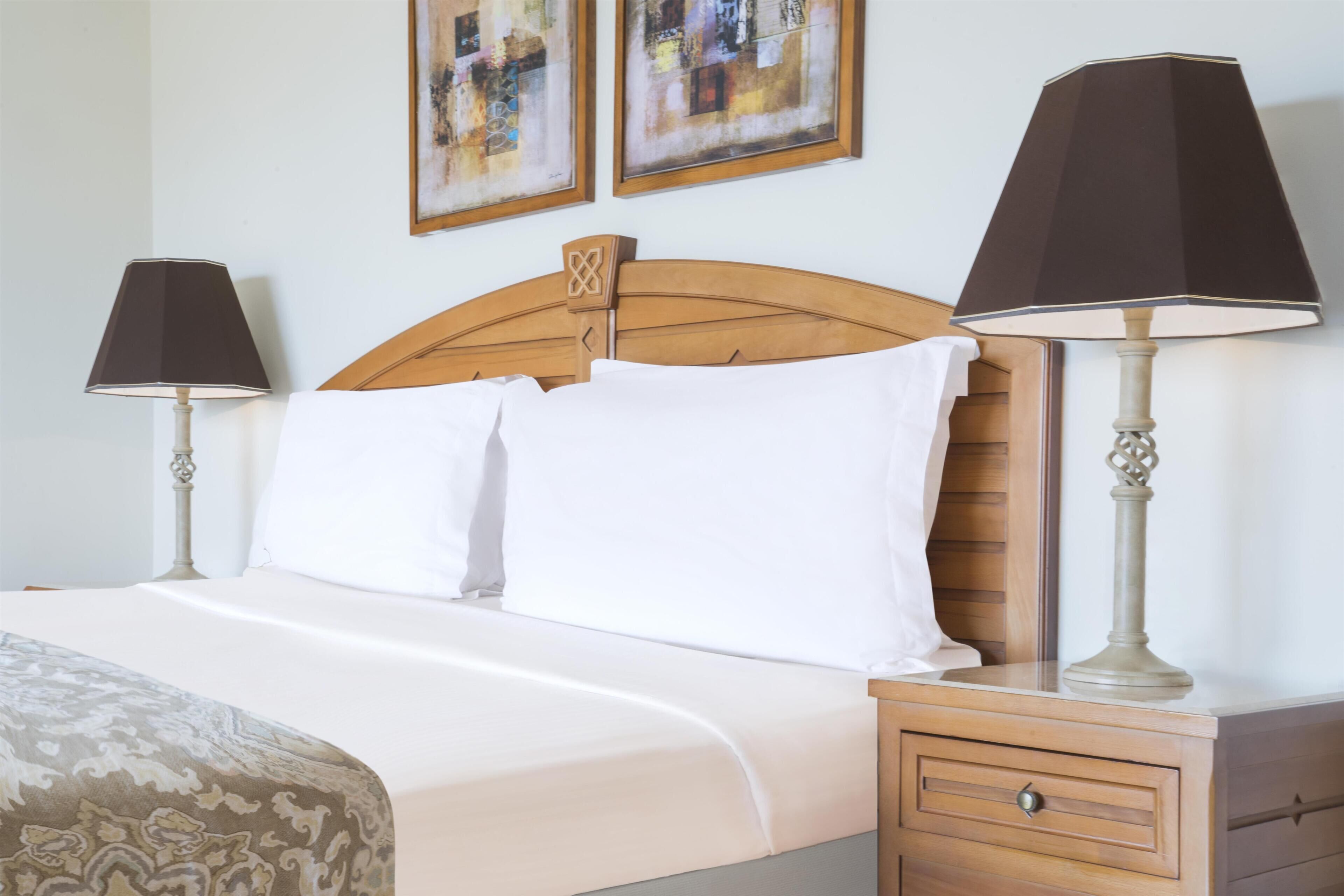 Una cama bien hecha con sábanas blancas, cabecera de madera y dos lámparas de noche.
