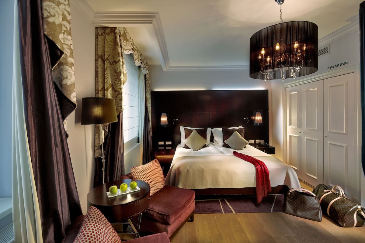 A hotel room with a bed, bedside tables, a desk, a chair, a lamp, a chandelier, and luggage.