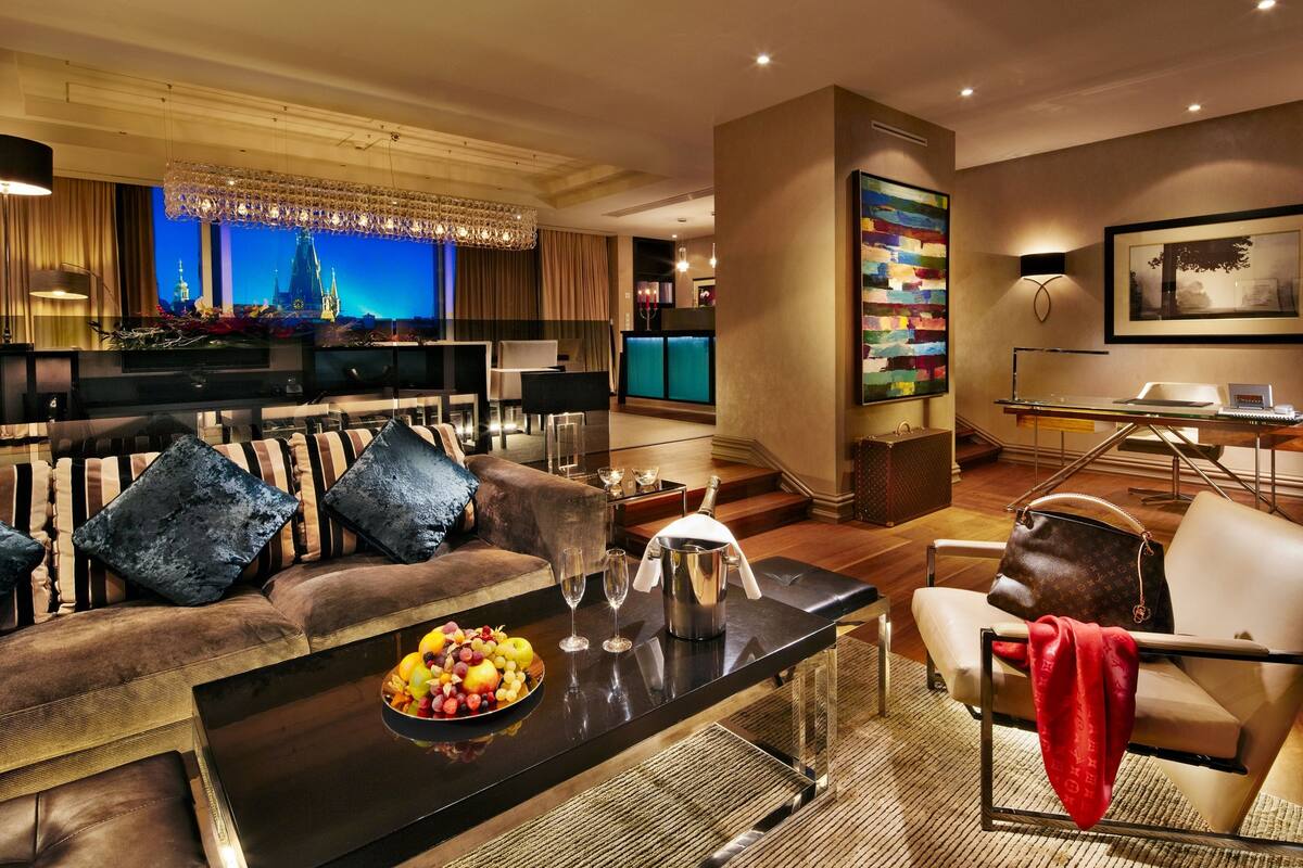 A modern living room with a sofa, armchairs, a coffee table with fruit and glasses, a TV, and a cityscape view.