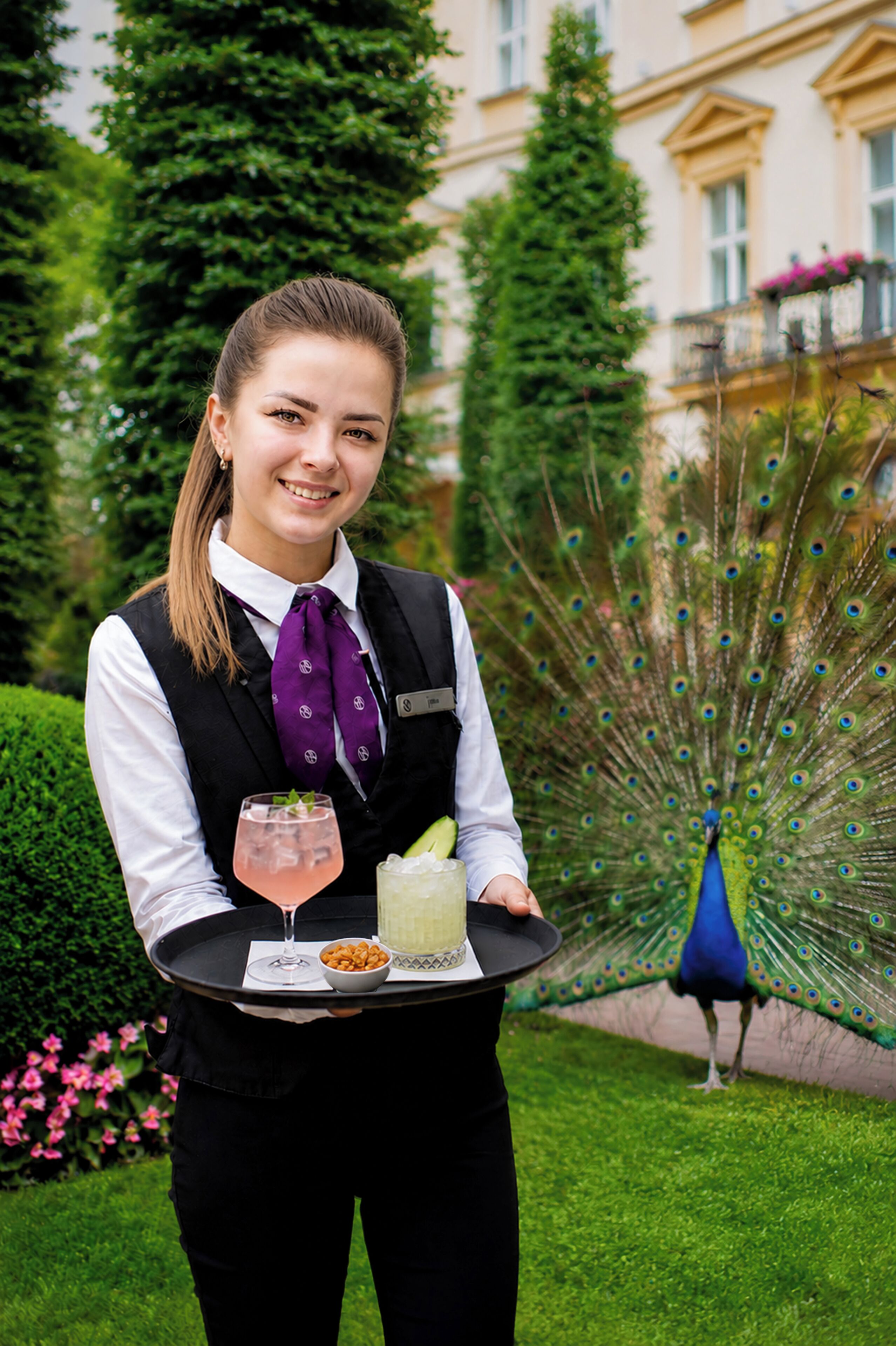 A person holding a tray with a drink and snacks, a peacock in the background, and a building with balconies.