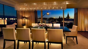 A dining area with a table set for six, surrounded by chairs, a chandelier, and a view of a cityscape at dusk. - The Grand Mark Prague - The Leading Hotels of the World (Prague)