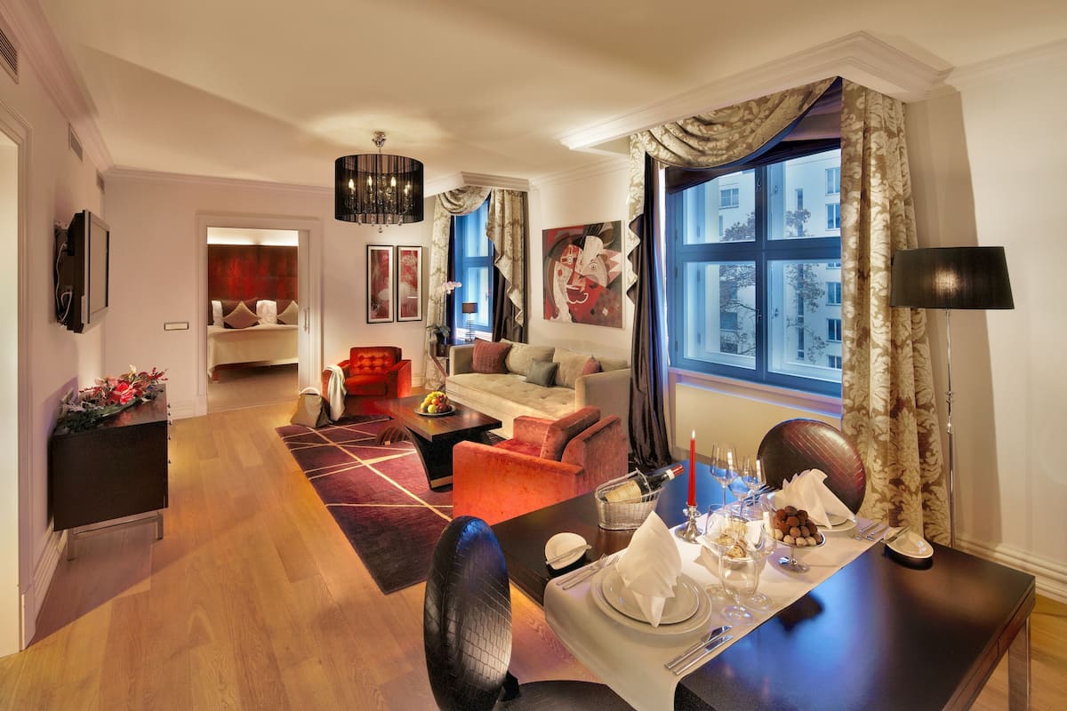 A modern living room with a sofa, armchairs, and a dining table set for a meal. A large window with curtains is visible, and a cityscape can be seen through the window.