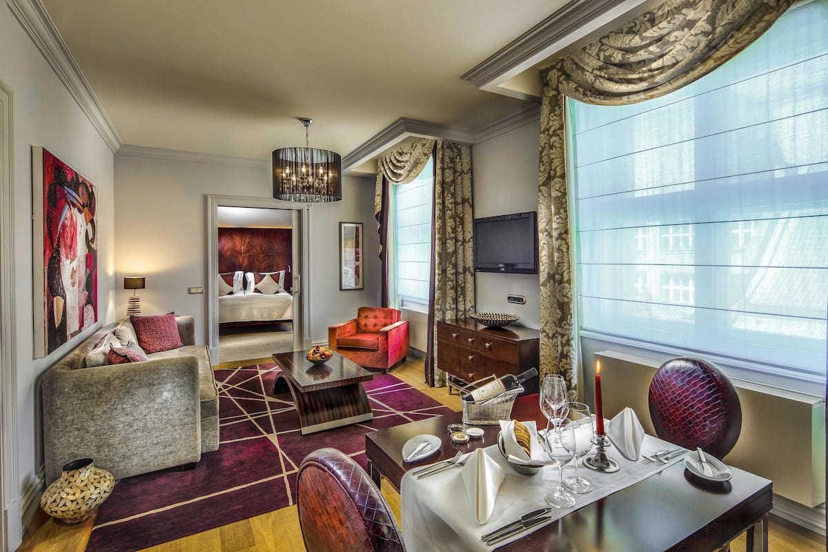 A spacious living room with a sofa, a red armchair, a dining table set for two, and a large window with curtains.