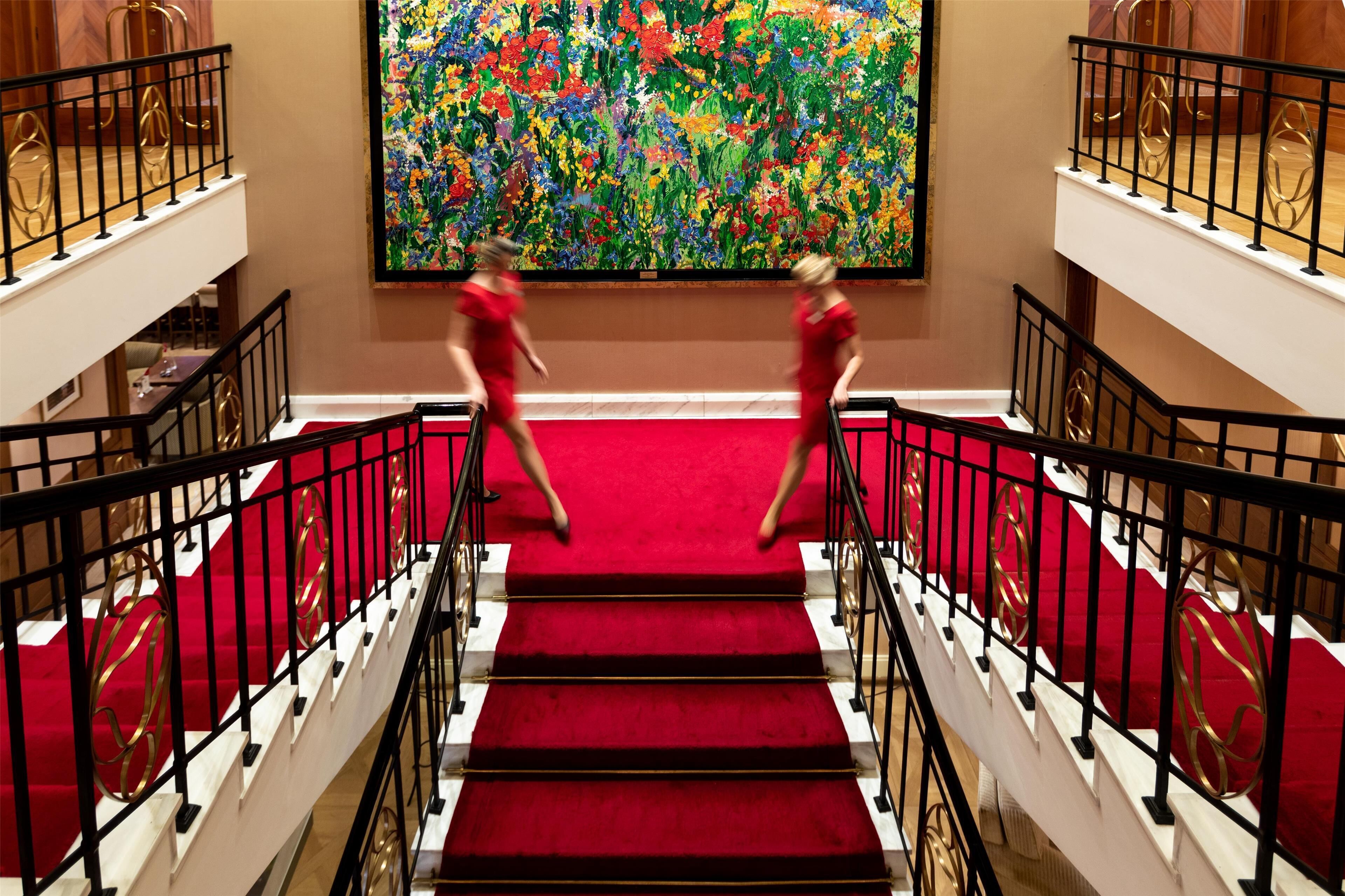 A grand staircase with red carpet and gold accents, leading to a room with a large abstract painting on the wall.