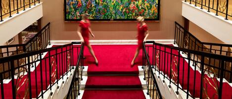 A grand staircase with red carpet and gold accents, leading to a room with a large abstract painting on the wall.