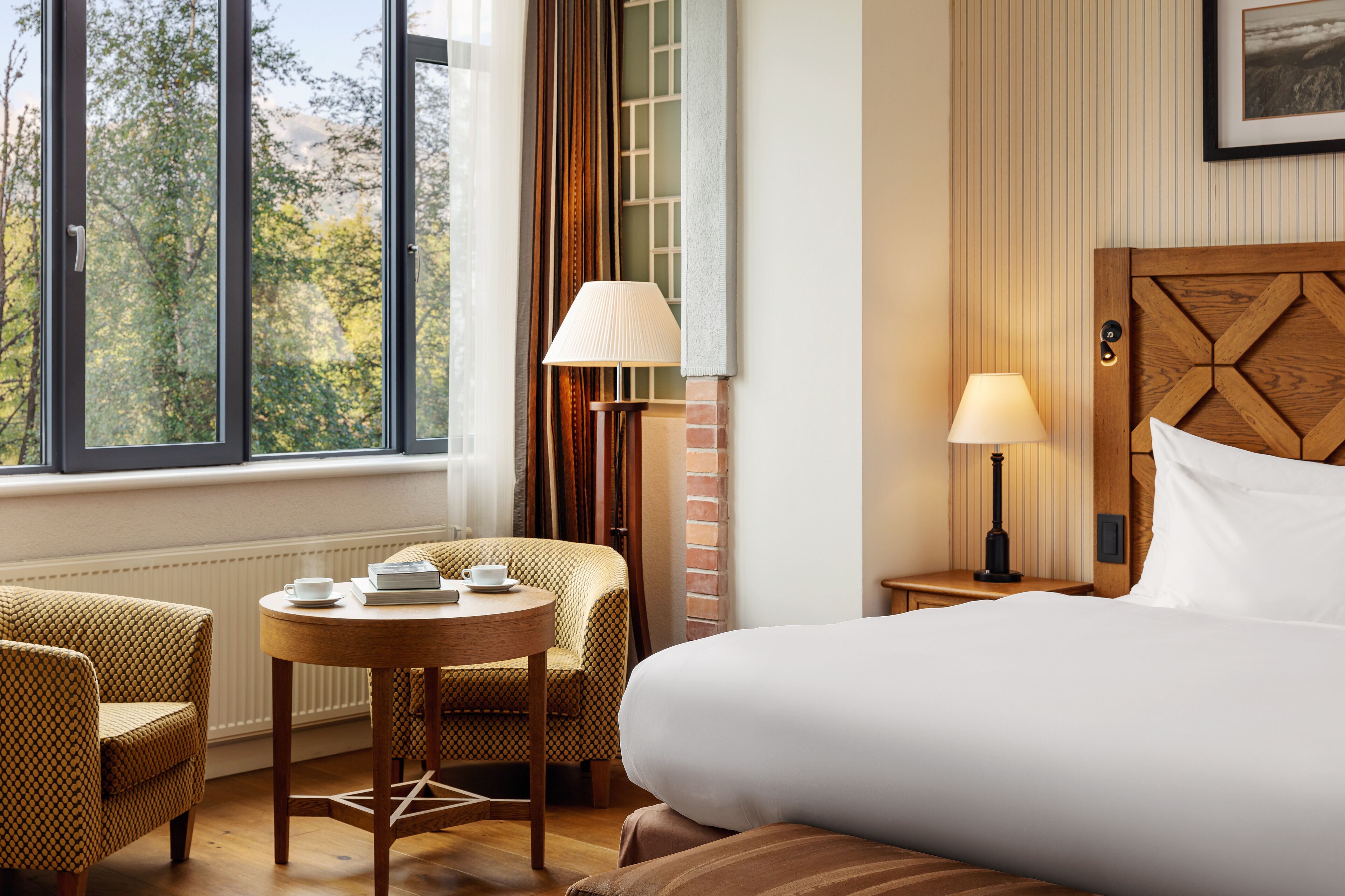 Ein Hotelzimmer mit einem großen Bett, einem runden Tisch mit Stühlen, einem Fenster mit Vorhängen und einem hölzernen Kopfteil.