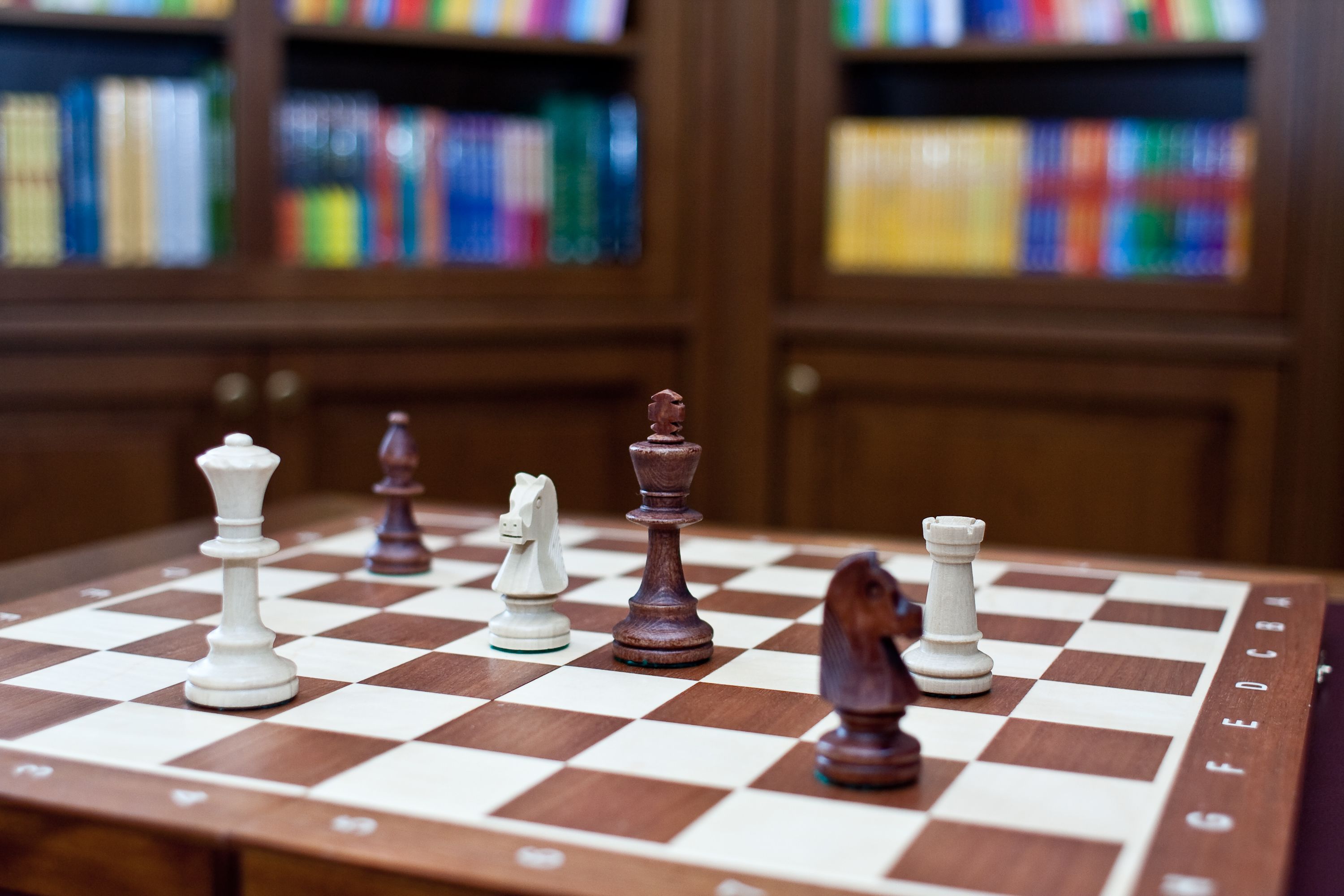 A chessboard with pieces on a wooden table, with bookshelves in the background.