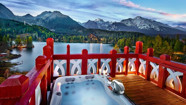 A wooden deck with a hot tub, a red railing, and a view of a lake and mountains.