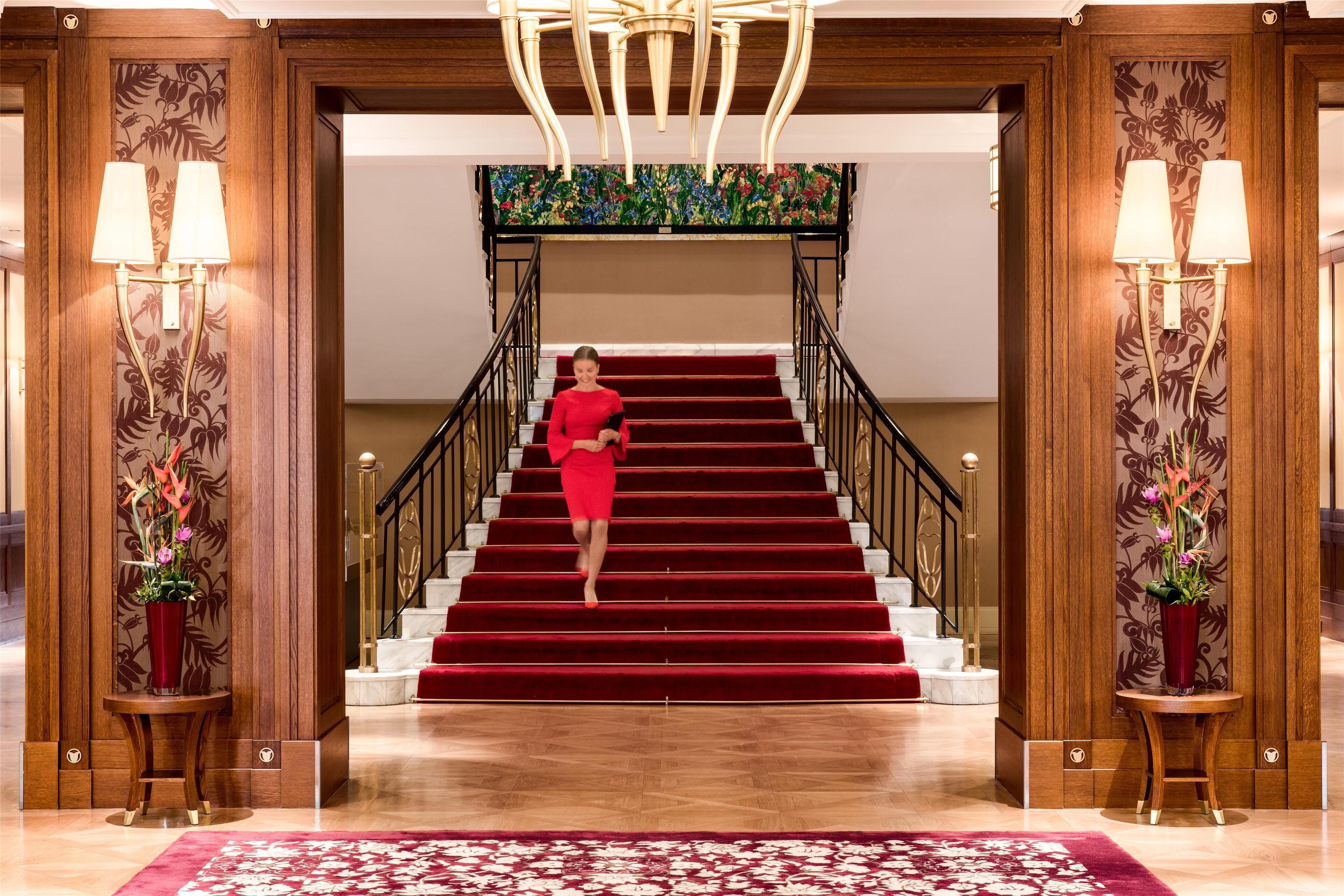 A woman in a red dress descending a red carpeted staircase in a hotel lobby with wooden paneling and two wall-mounted lamps.