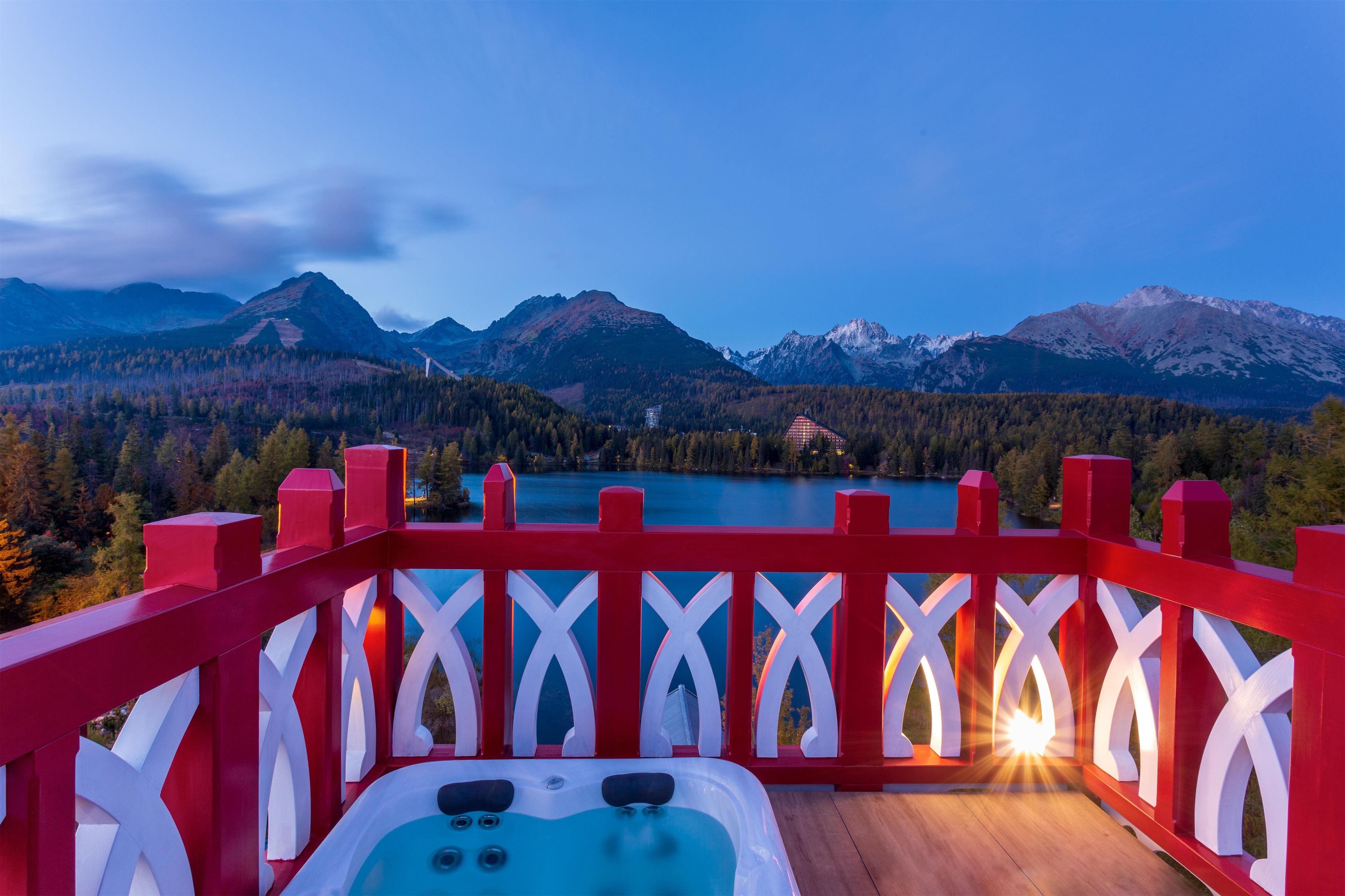 A wooden deck with a red railing, overlooking a lake and mountains.