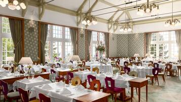 A formal dining room with multiple tables set for service, large windows, and classic chandeliers.