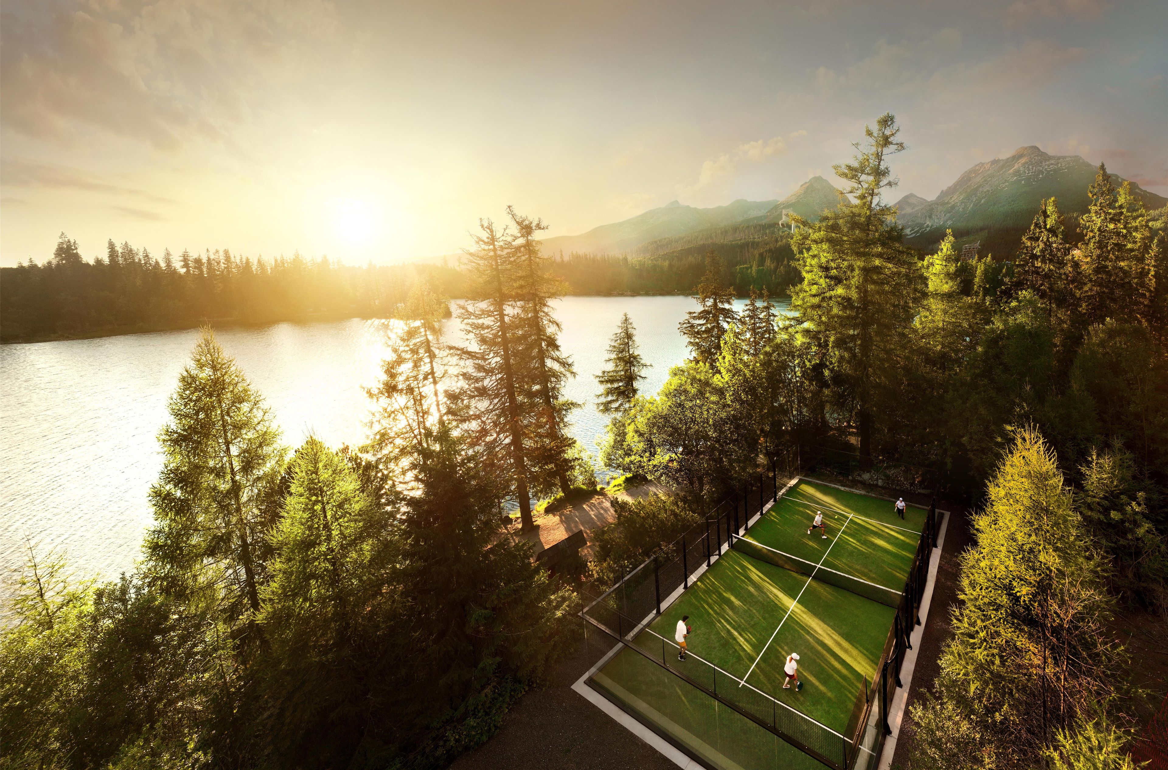 A tennis court surrounded by trees and a lake with mountains in the background.