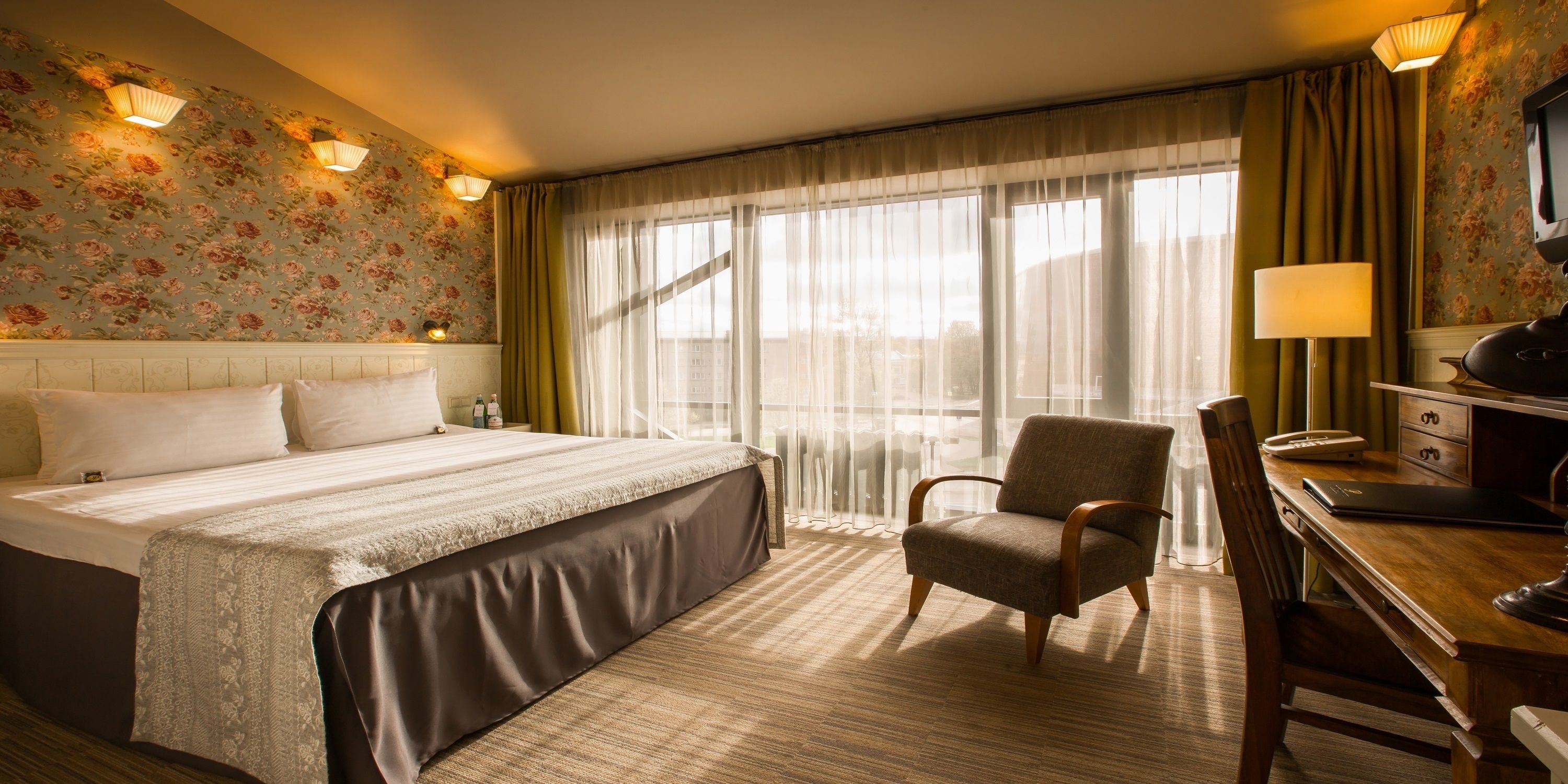 A hotel room with a large bed, a desk with a chair, a television, and a window with sheer curtains.