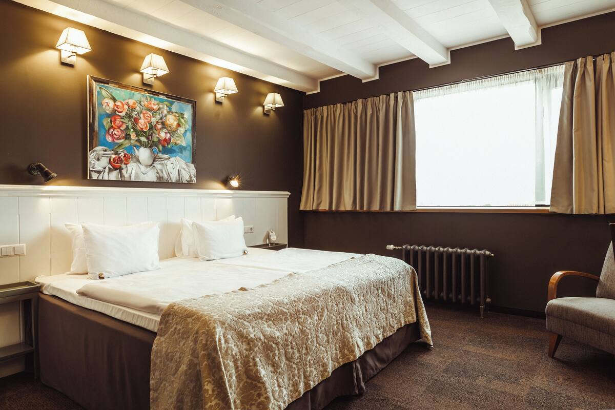A hotel room with a large bed, two wall-mounted lamps, a radiator, a window with curtains, and a painting on the wall.