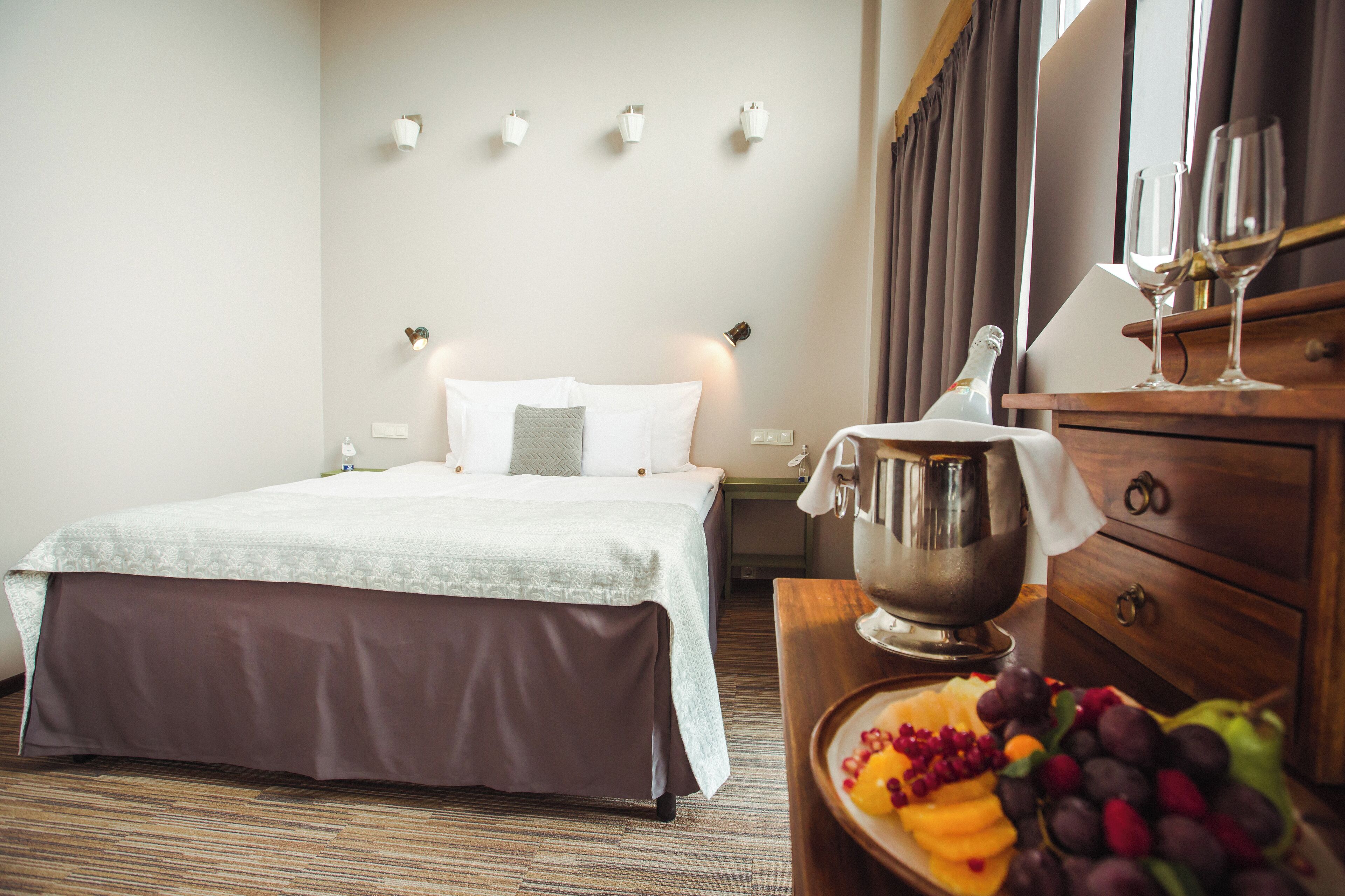 A hotel room with a neatly made bed, a wooden dresser with a bottle and glasses, and a fruit platter on the dresser.