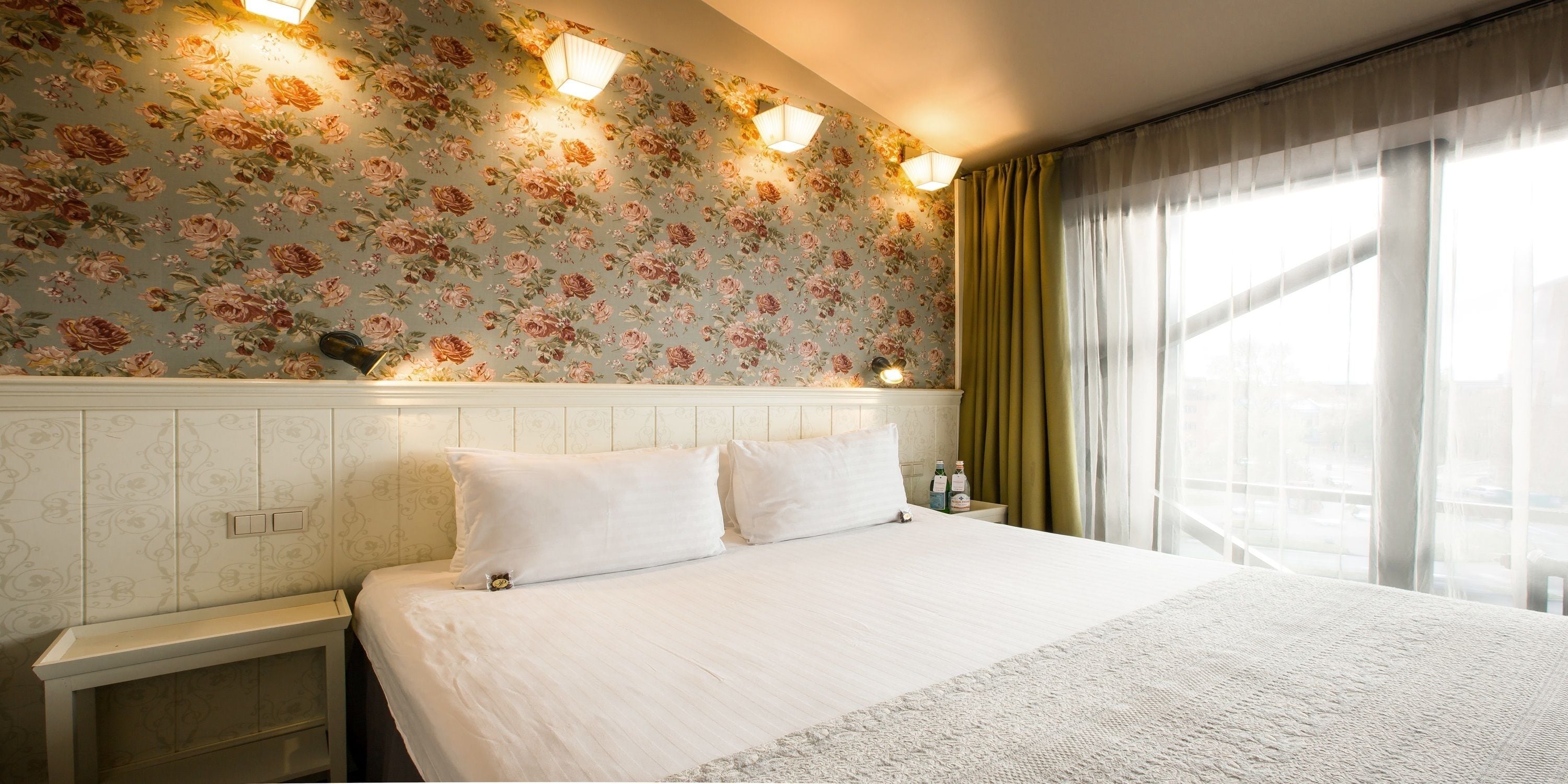 Een hotelkamer met bloemenbehang, een bed met witte lakens, een nachtkastje met een lamp en een raam met gordijnen.