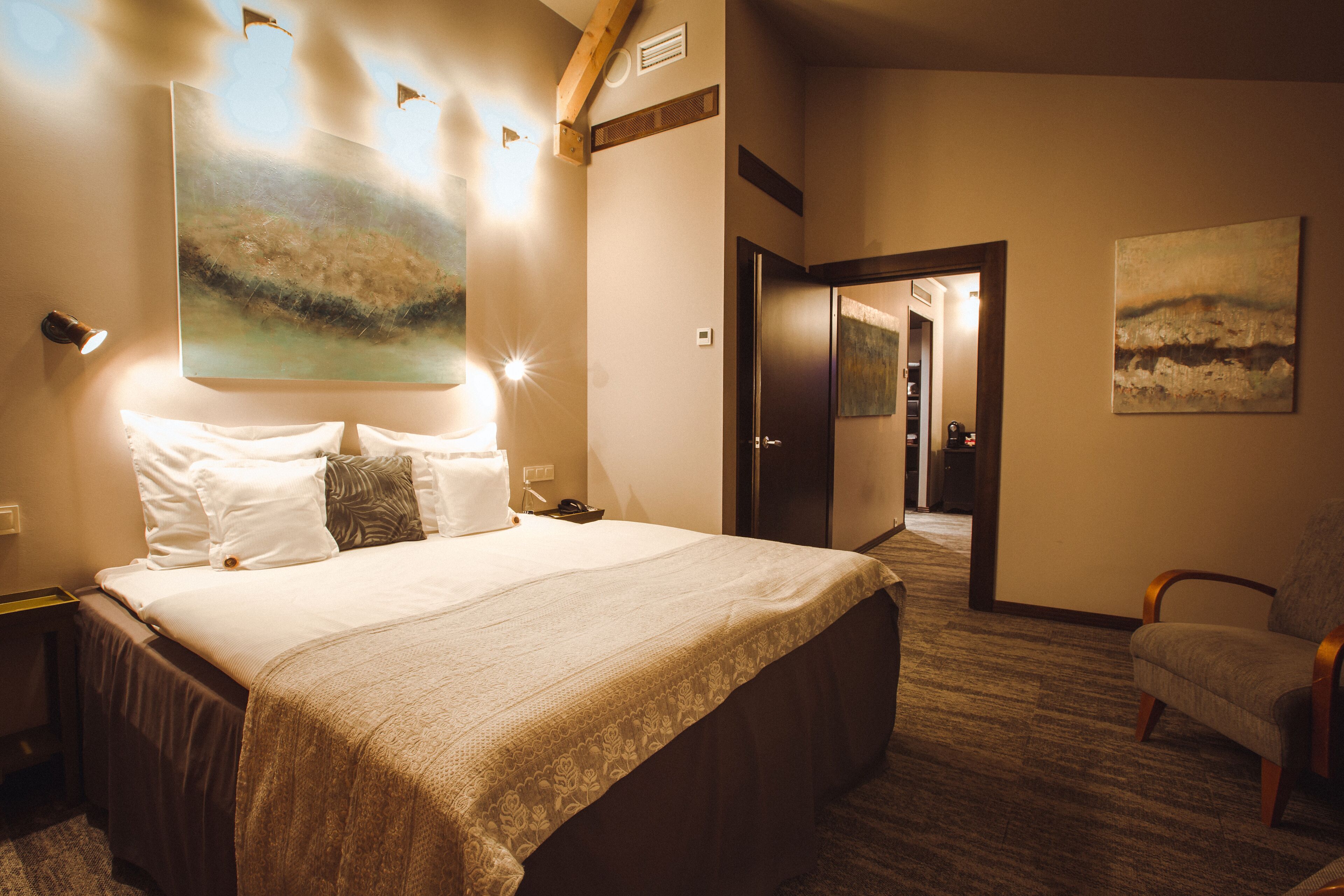 Une chambre d’hôtel avec un grand lit, une chaise, un tableau au mur et une porte donnant sur une autre pièce.