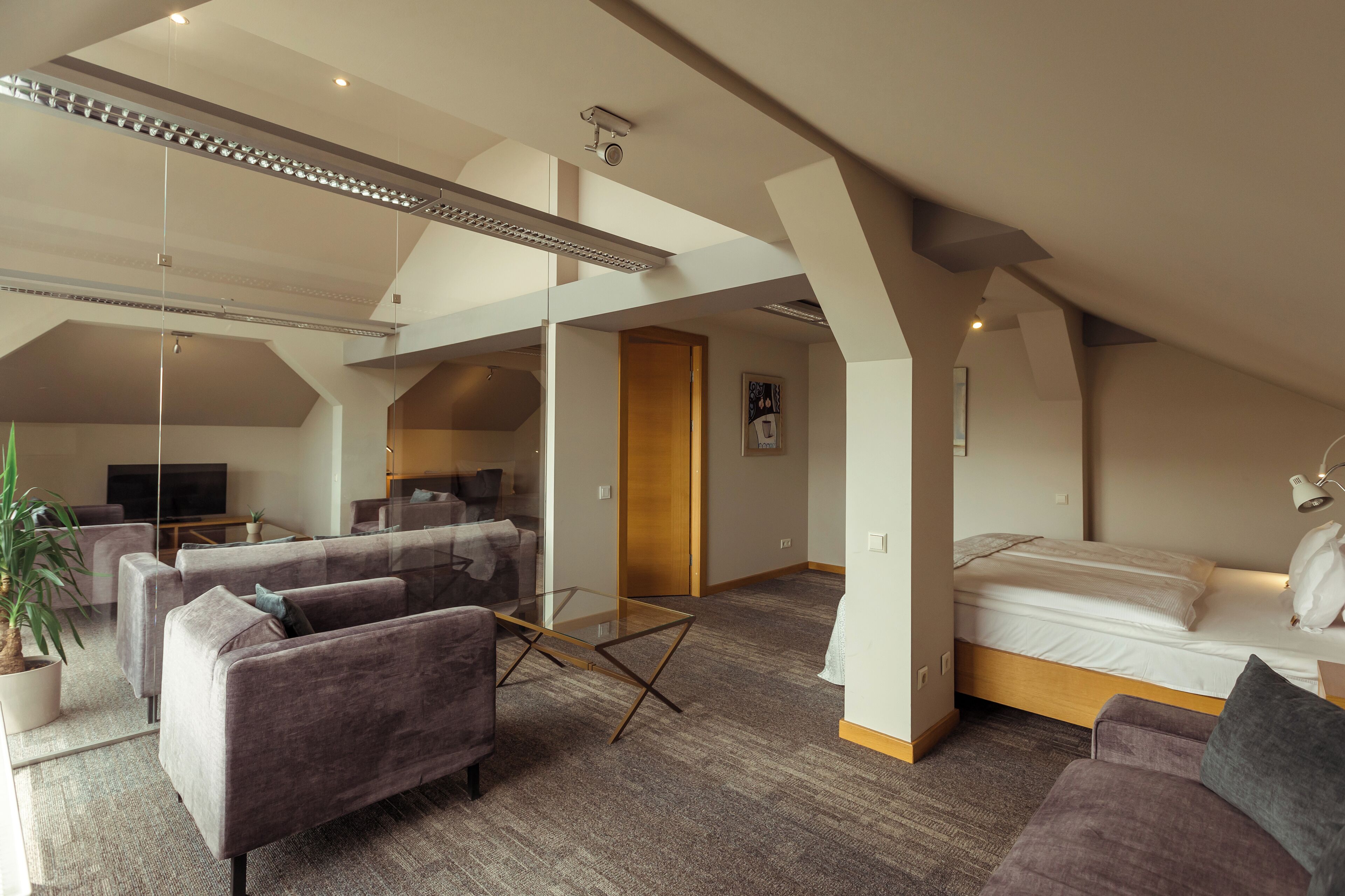 A modern loft-style room with a high ceiling, a glass partition separating the living area from the bedroom, and a large window providing natural light.
