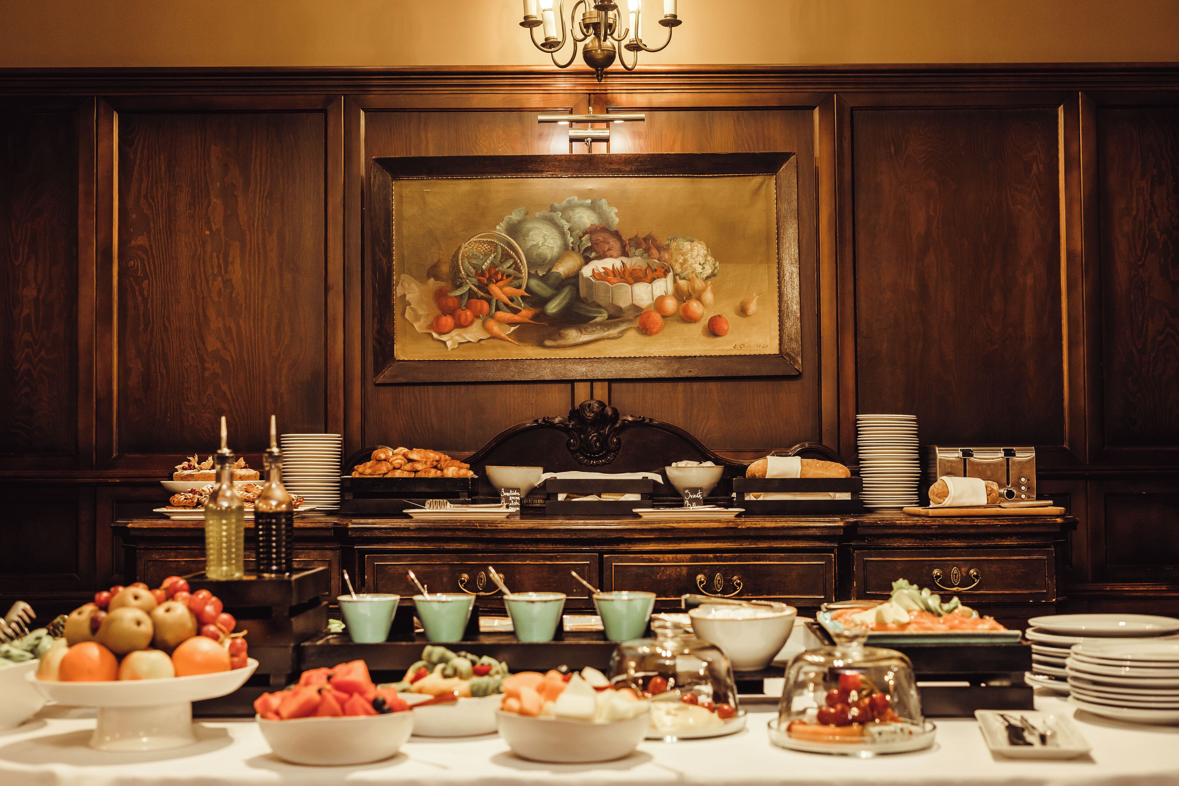 A buffet table with various food items and dishes, set against a backdrop of wooden paneling and a large painting.