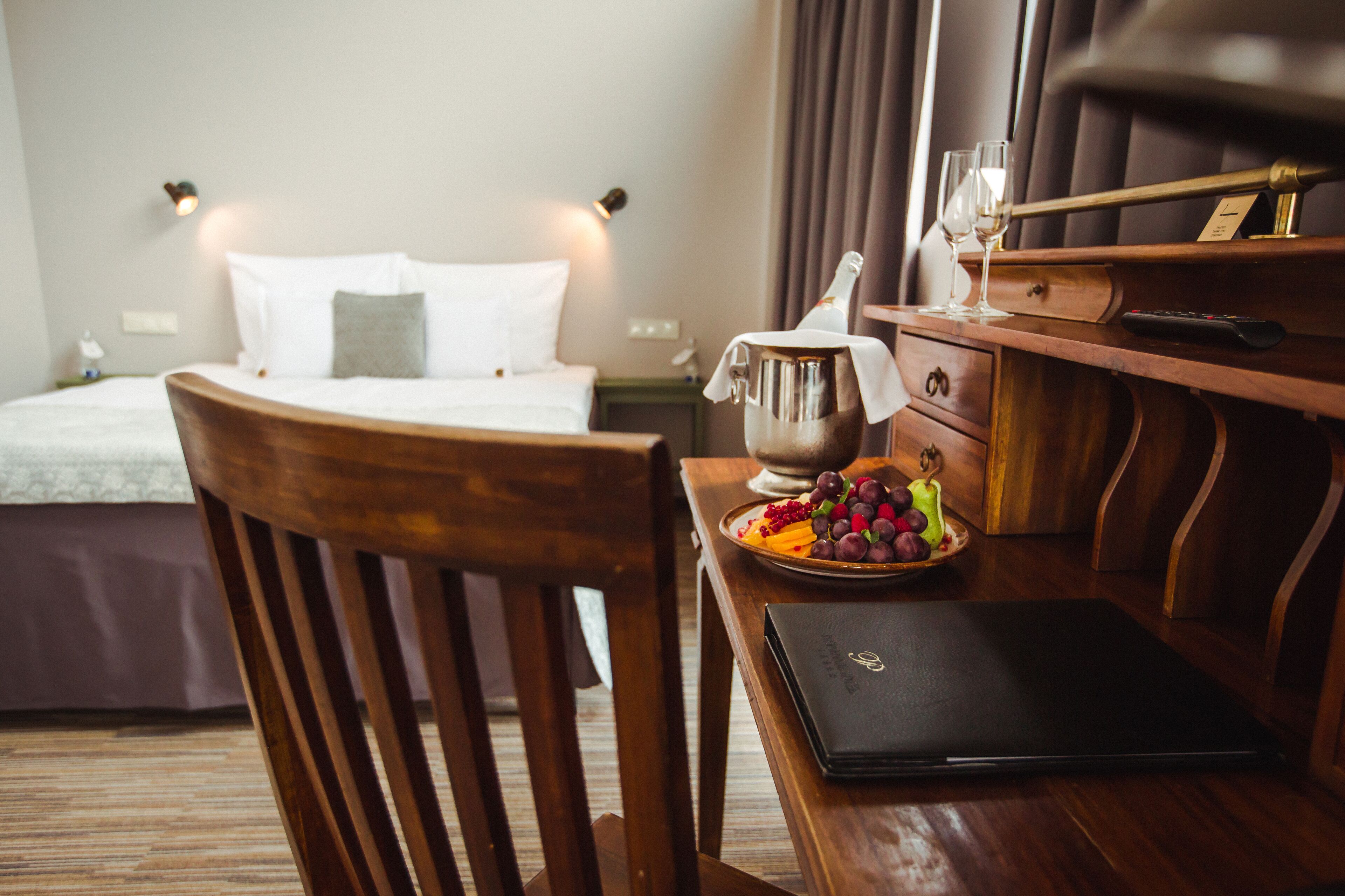 A hotel room with a bed, bedside table with a bottle, glasses, and a fruit plate.